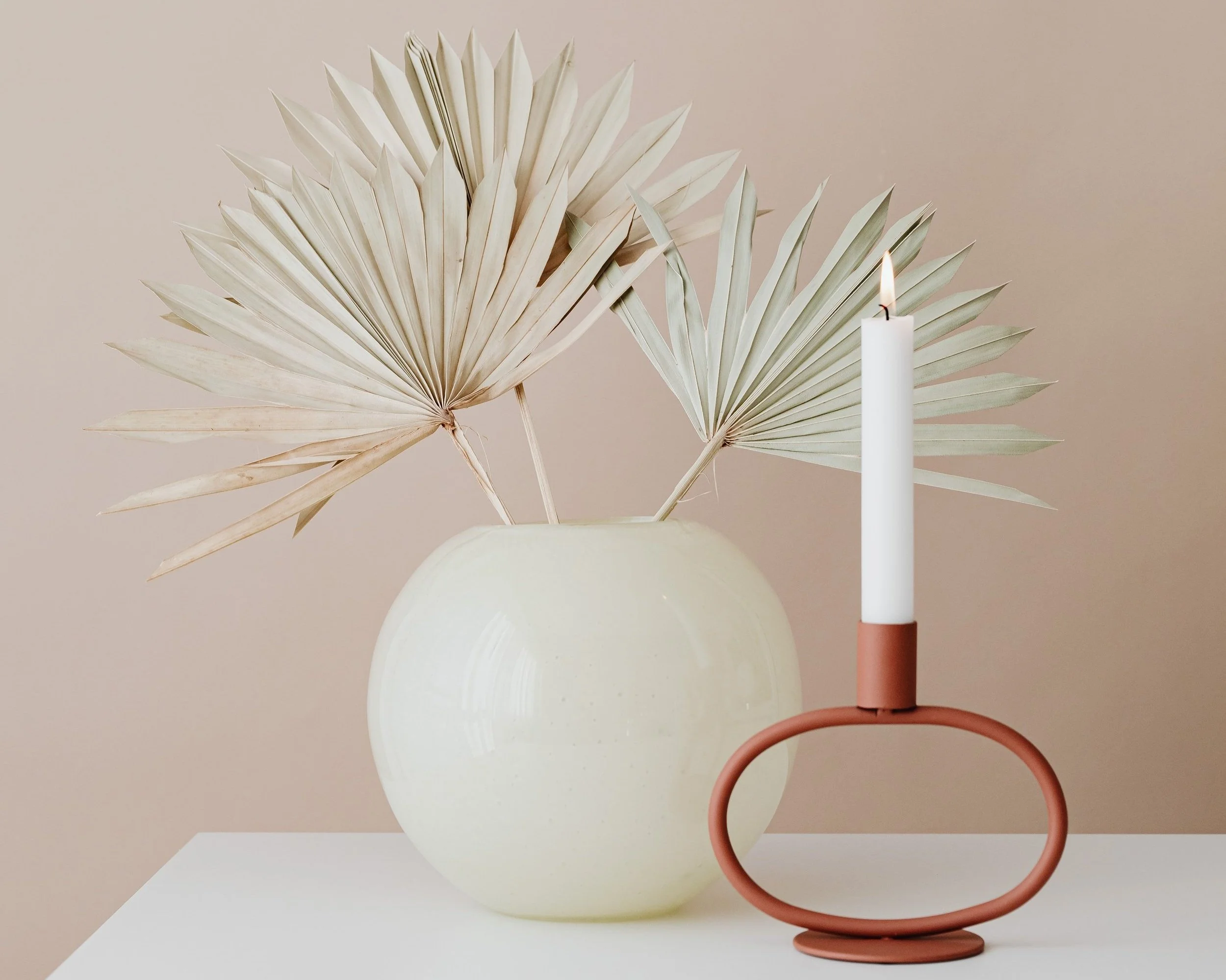 Dried fan palm leaves in a tan ceramic vase placed next to a terracotta candle holder with a white pillar candle.