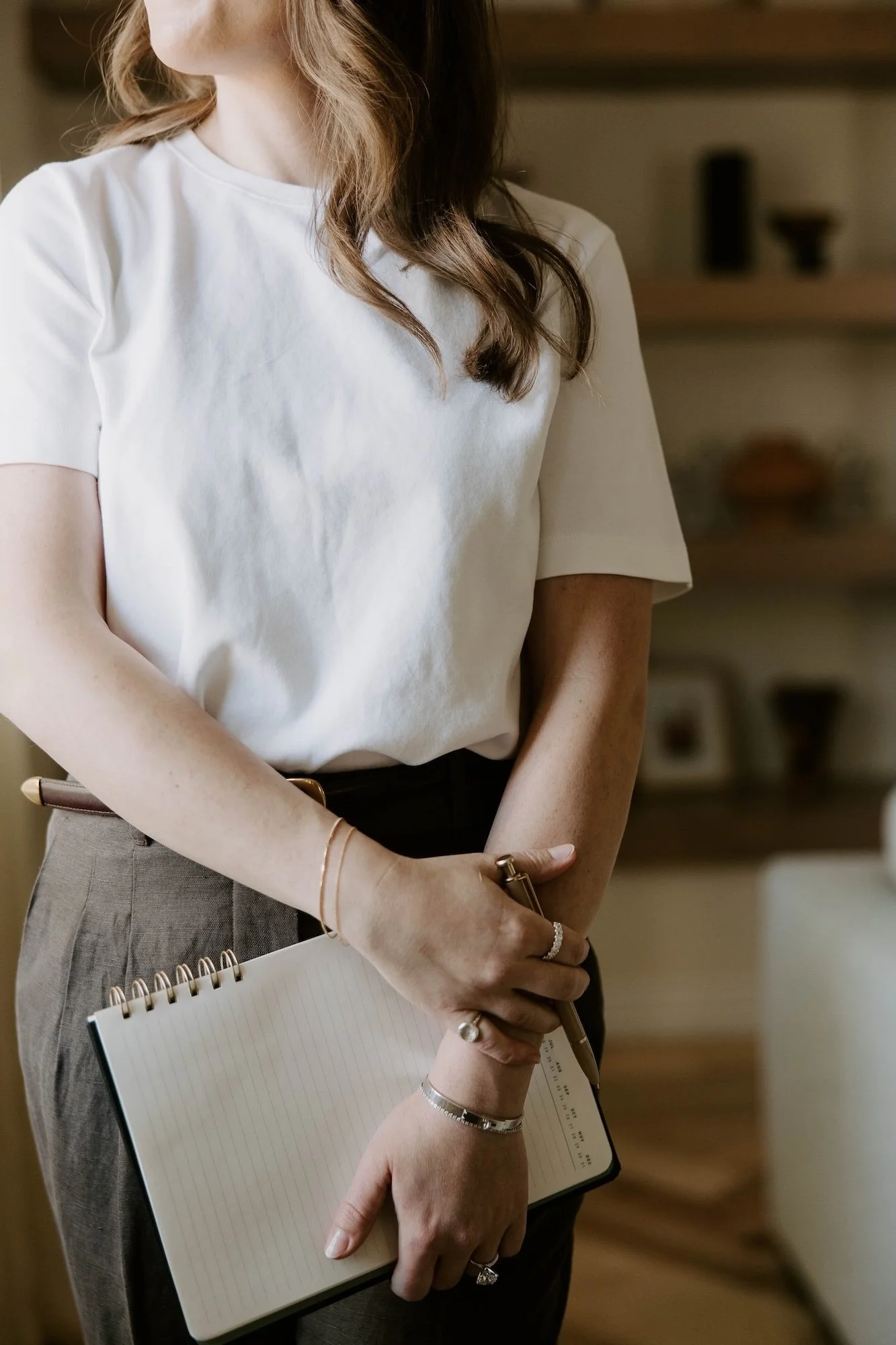 A female therapist standing thoughtfully near natural light while holding a open notebook in a warm office