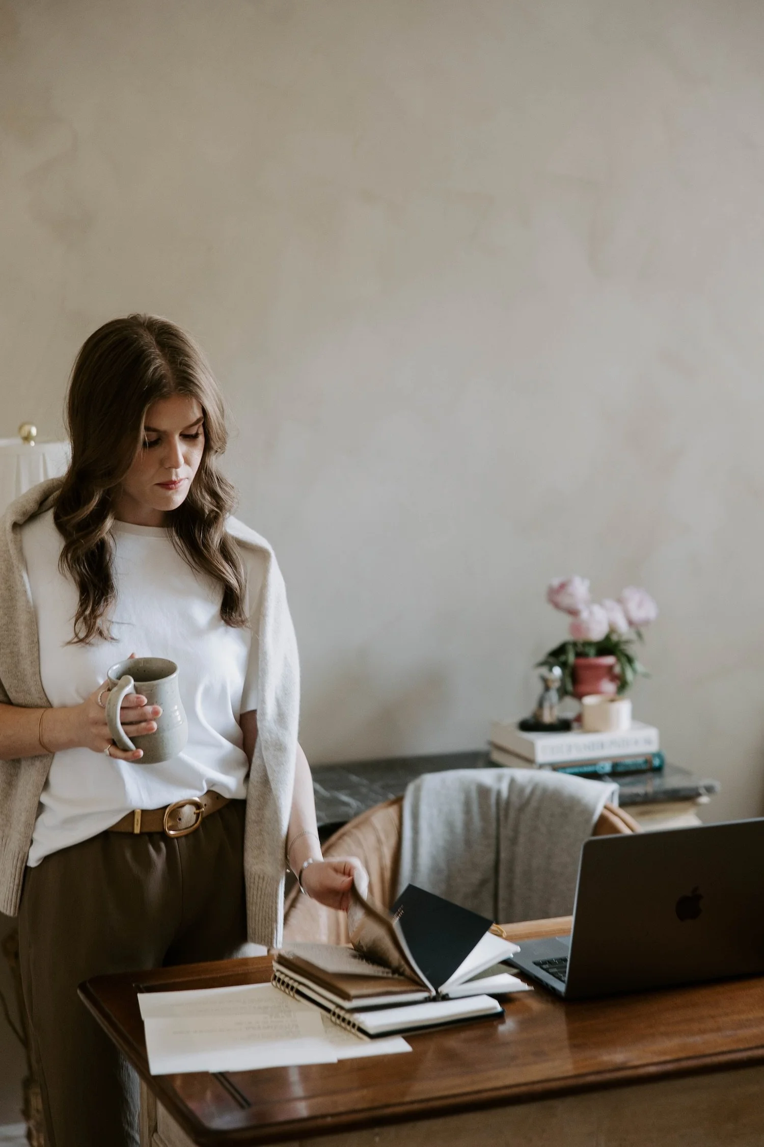 A woman with wavy brown hair is holding a gray mug and looking down. She is dressed in a white shirt and tan pants with a brown belt. She is in a room with a table that has notebooks and a laptop on it. In the background, there is a shelf with books, a small plant, and decorative items.