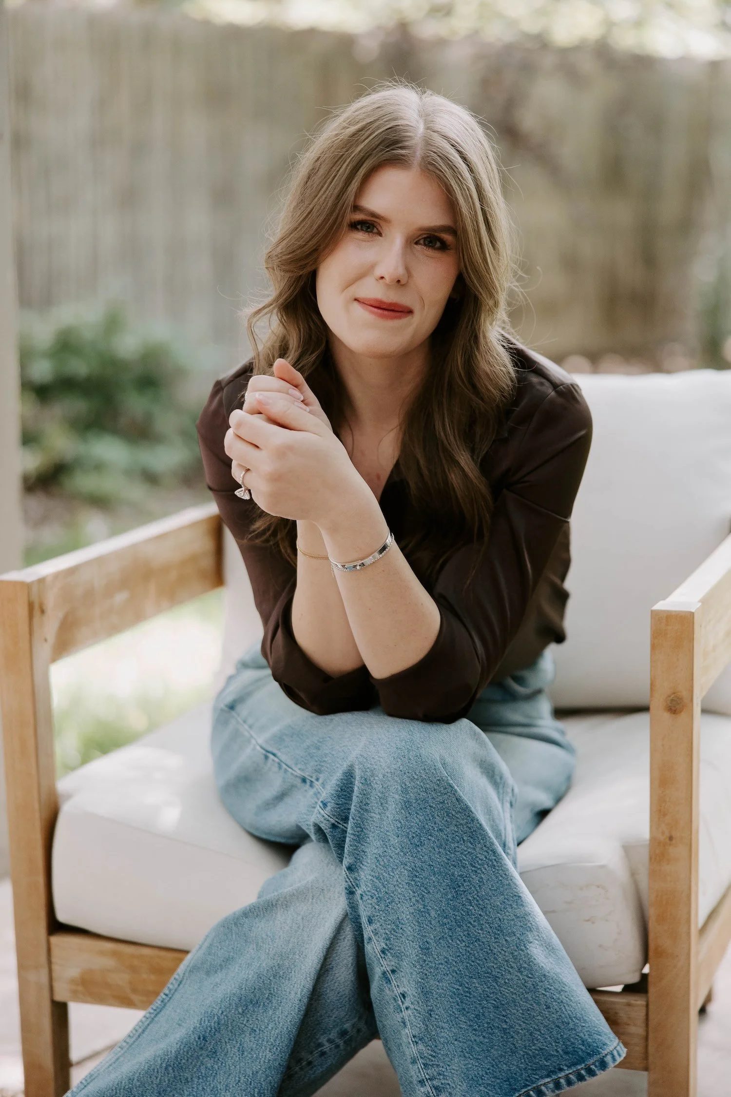 A woman with long wavy brown hair sitting on a white sofa, wearing a dark brown top and light blue jeans, smiling and looking at the camera, with a light-colored outdoor background.