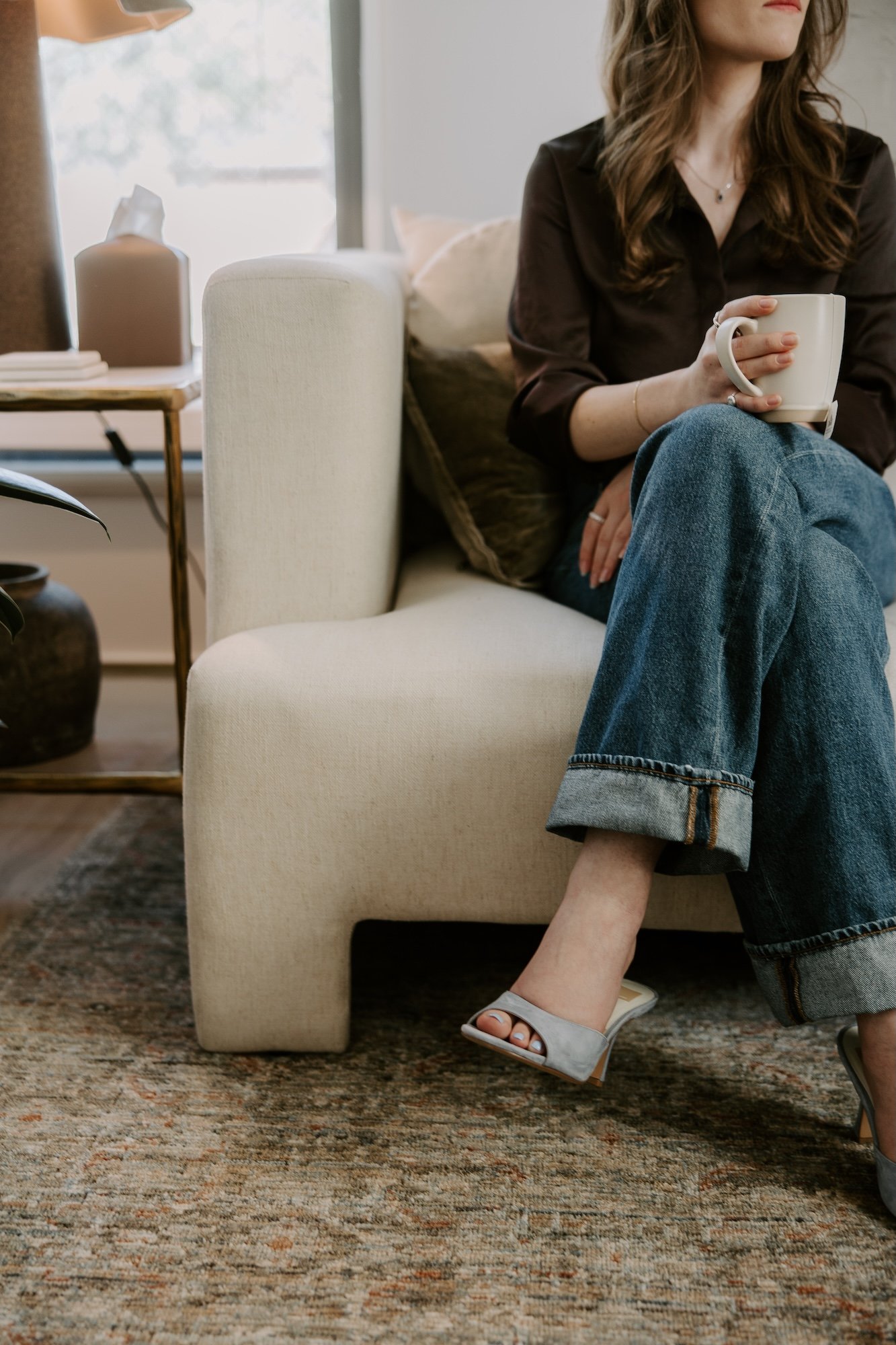 A woman sitting on a white upholstered sofa, holding a white mug, with a side table and decorative items in a cozy living room.