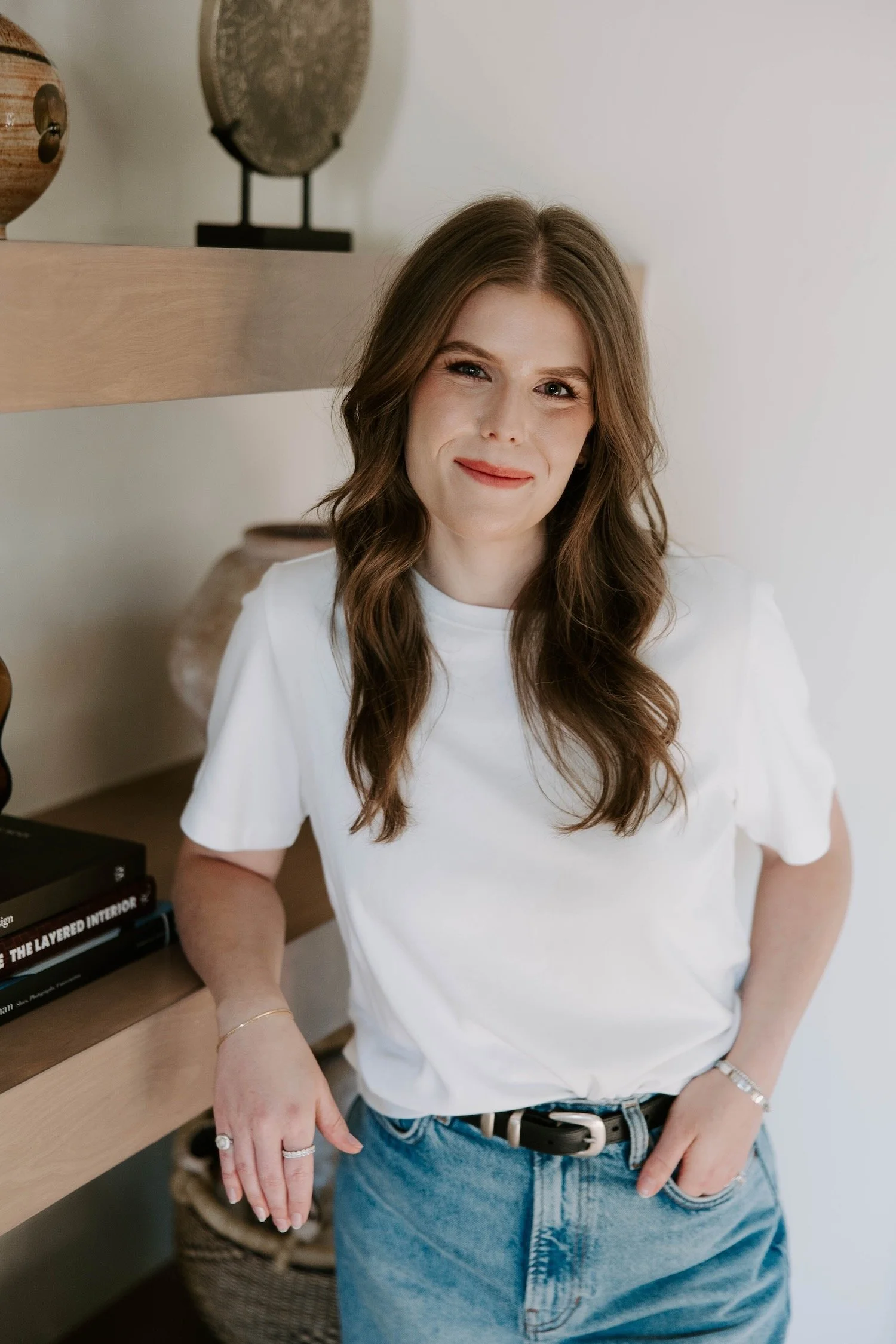 A woman with long brown hair, wearing a white t-shirt and blue jeans, standing indoors near a wooden shelf with decorative plates and books.