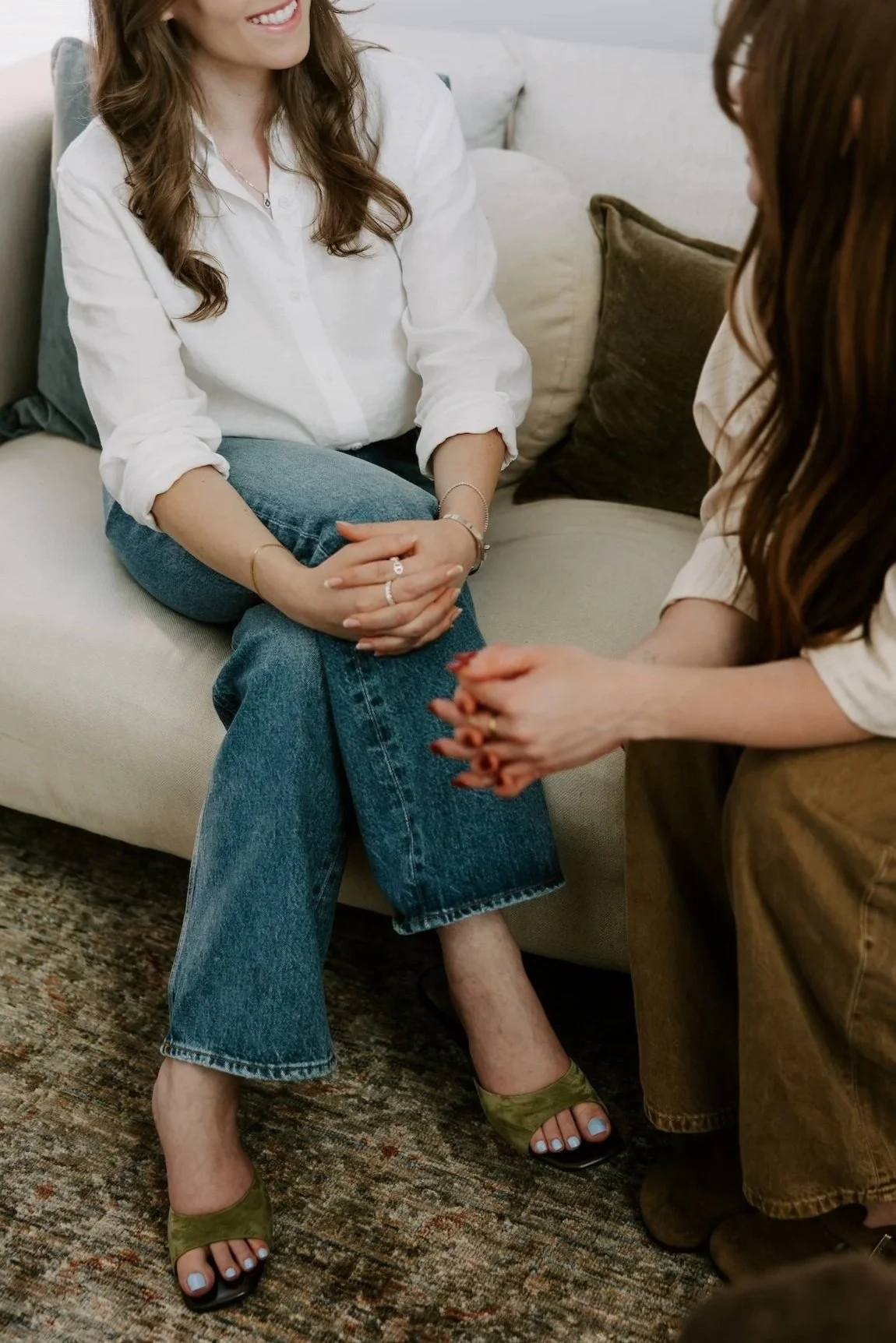 Two women sitting on a couch, engaged in conversation, with one woman smiling and the other with clasped hands.