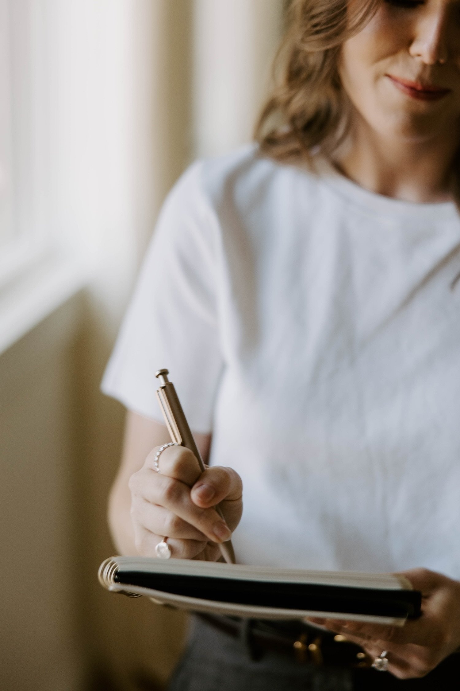 A woman writing in a notebook with a pen, wearing a white shirt and jewelry, with soft natural lighting.