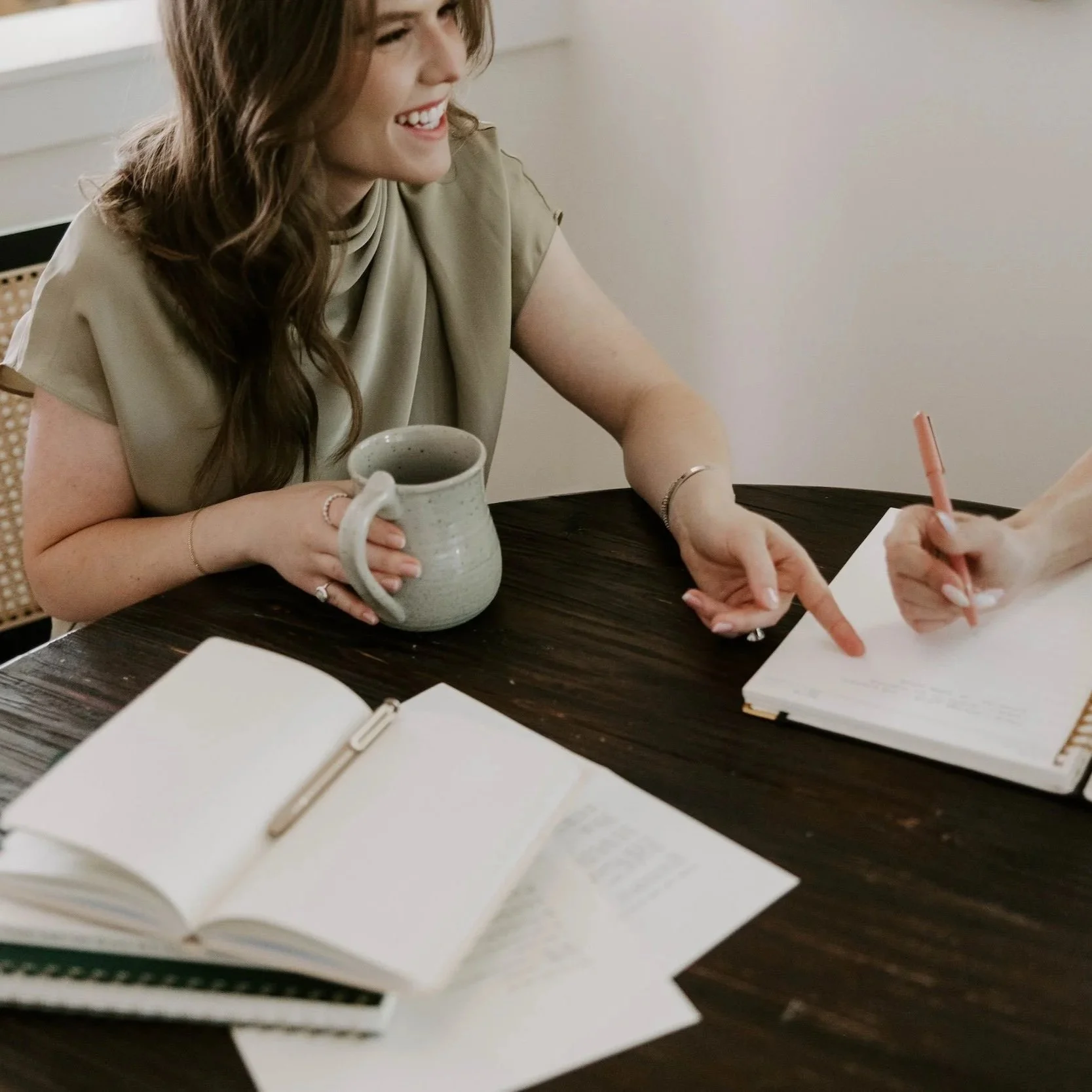 A woman with long, wavy brown hair laughing at a table, holding a ceramic mug filled with a beverage, with a notebook and pen in front of her, and another person writing in the notebook.