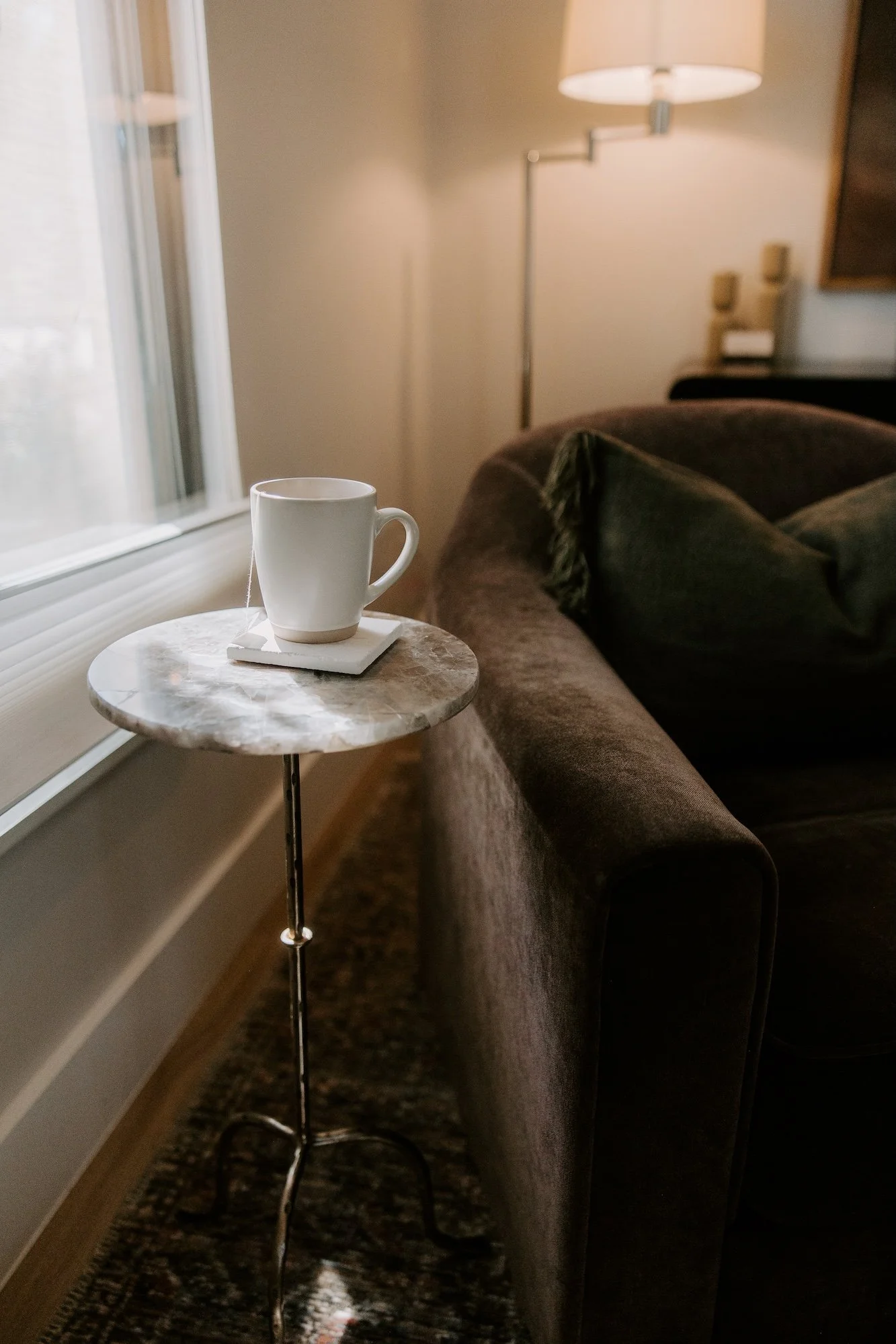 A white coffee mug on a small round marble-top table next to a brown sofa and a window with a no-tear closed blind, in a cozy room with a lamp and warm lighting.