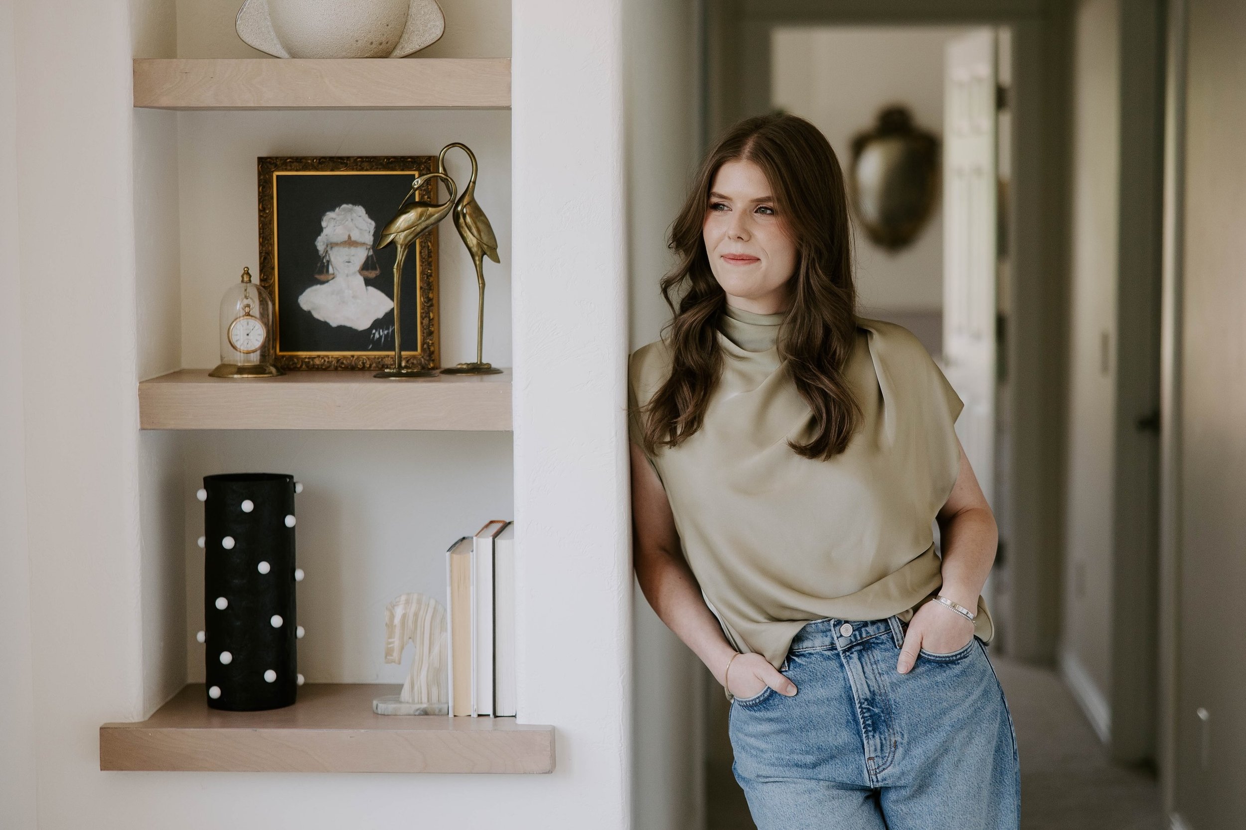 A woman with long brown hair wearing a beige top and jeans, standing in a hallway next to a bookshelf with decorative items and a framed drawing.
