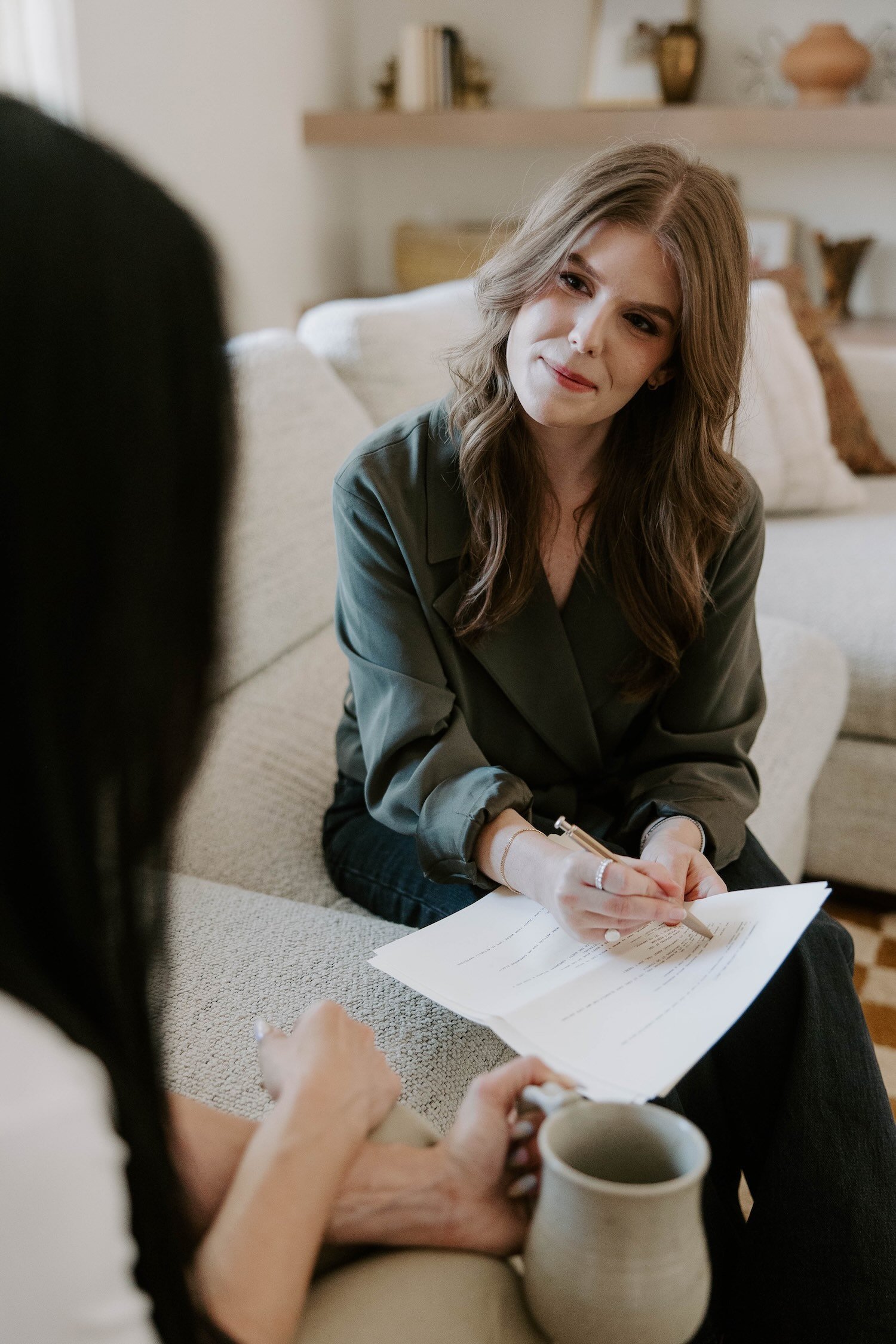 Two women sitting on a beige couch with coffee mugs, papers and a pen, having a conversation in a cozy living room.