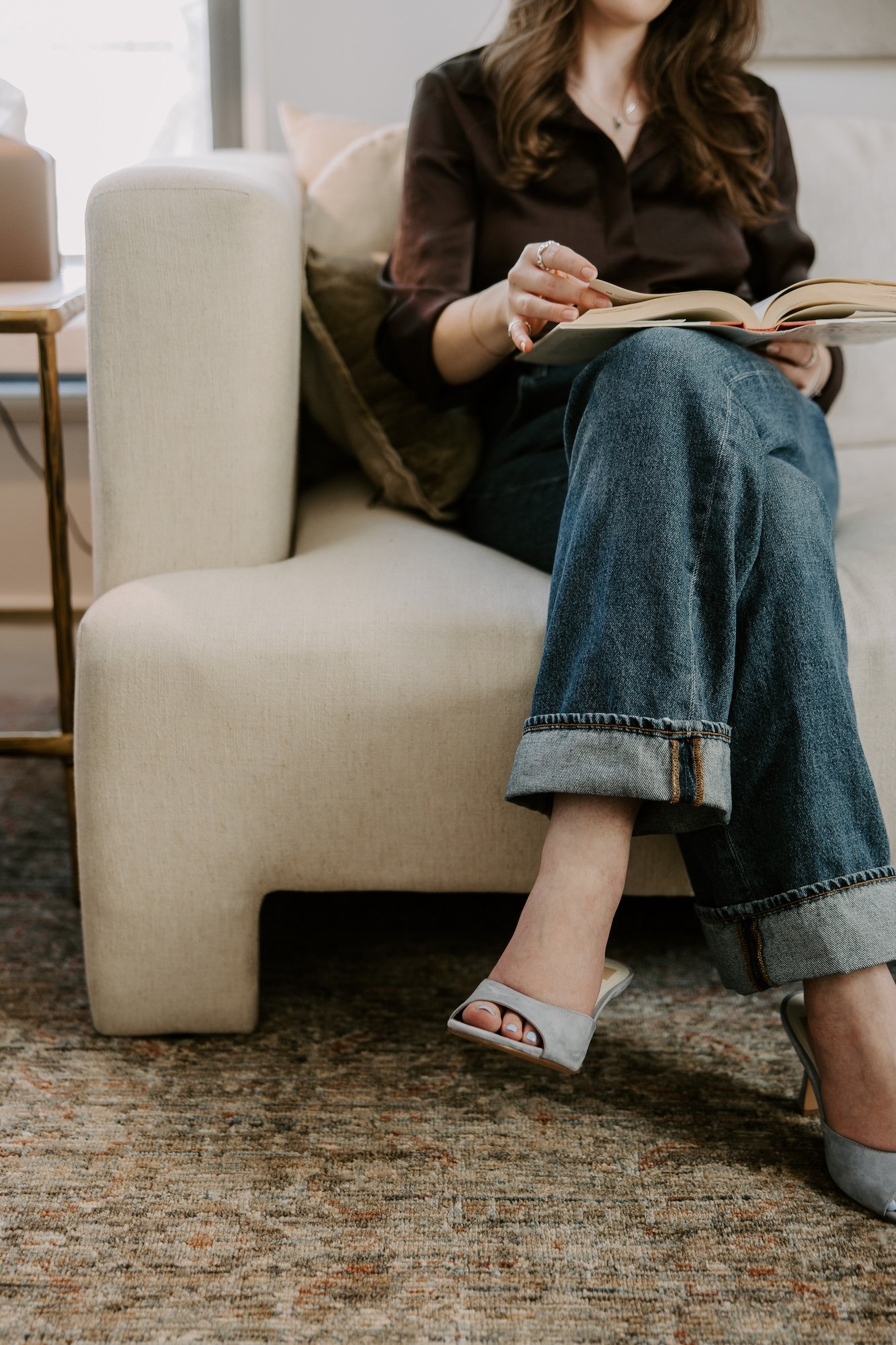 A woman sitting on a cream-colored sofa reading a book, wearing blue jeans and high heels, with her legs crossed at the ankle.