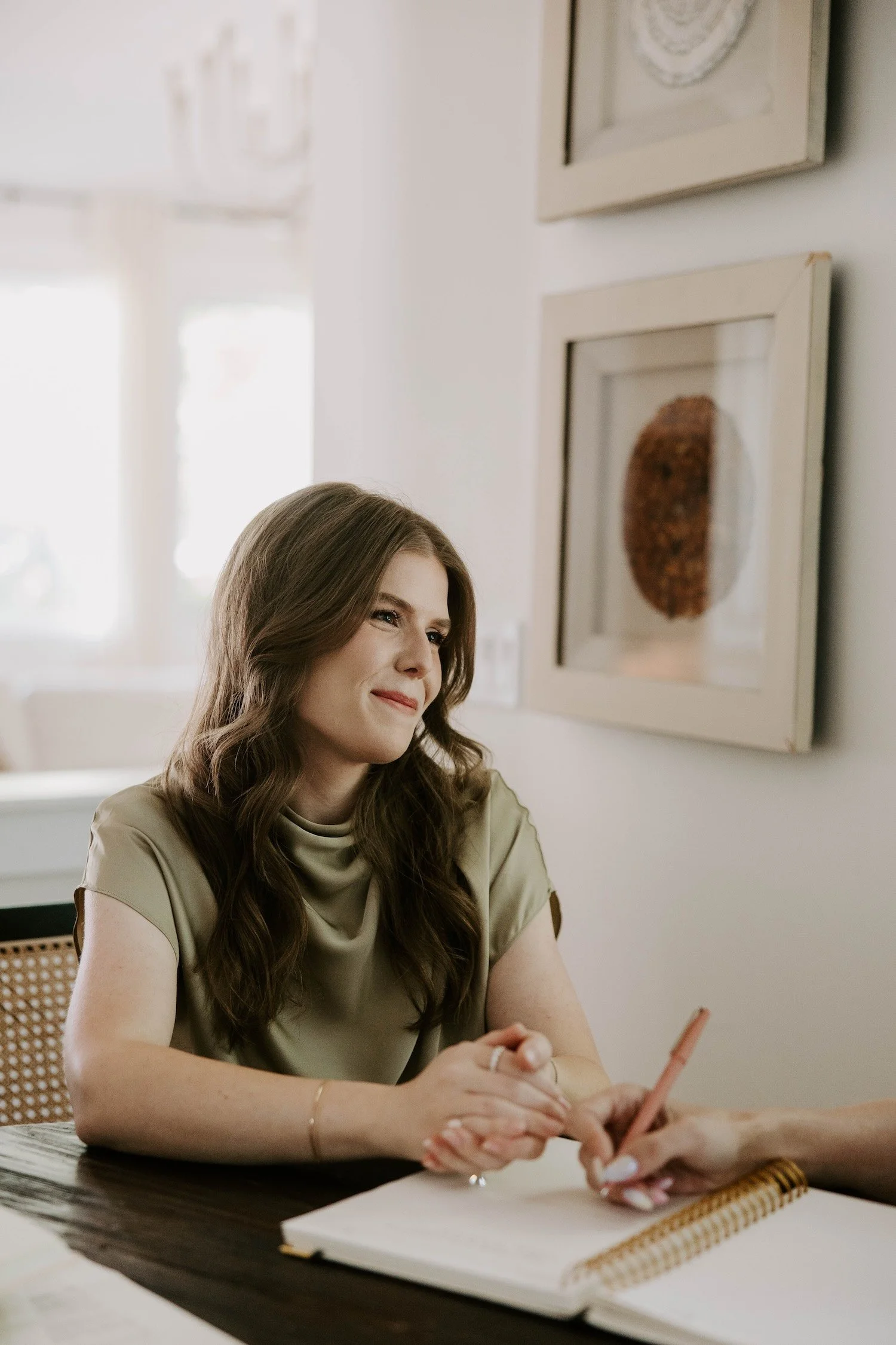 A woman with long wavy brown hair, wearing a beige blouse, sitting at a table, holding a pink pen, and smiling slightly, with framed artwork on light-colored walls behind her.