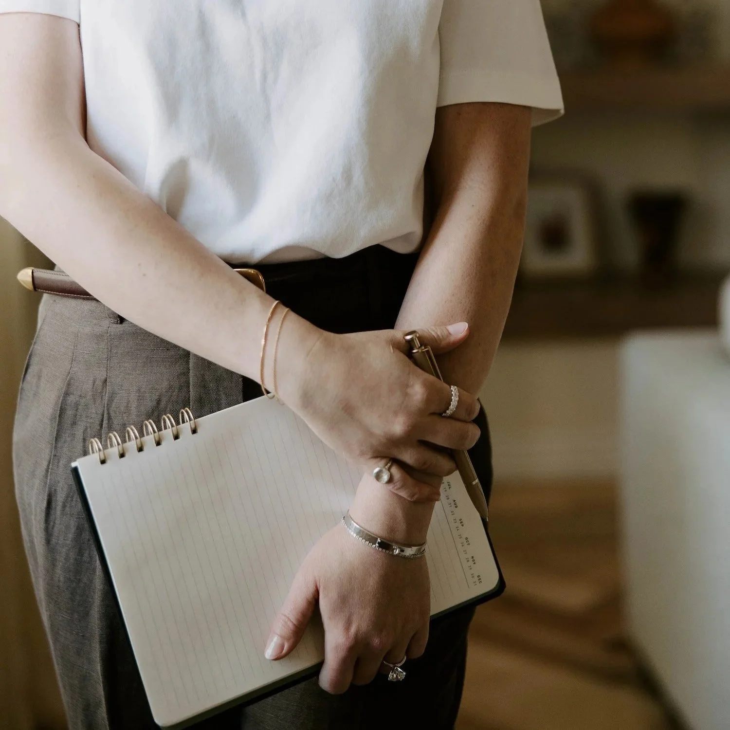 Close-up of a woman wearing a white T-shirt and gray pants, holding a notebook and a pen, standing in an indoor setting with blurred background shelves.