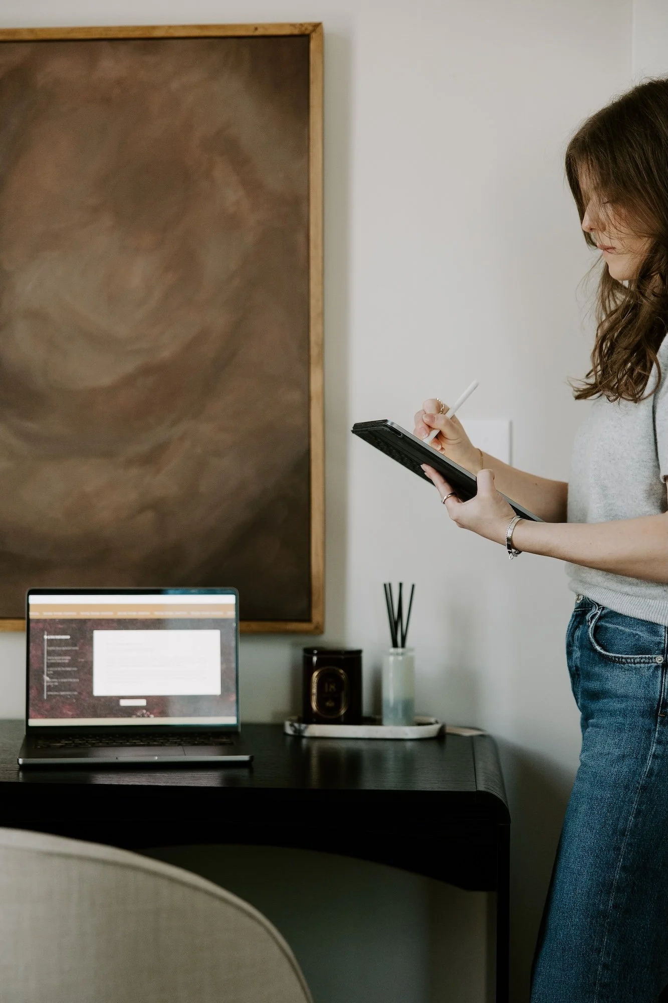 A woman with long brown hair wearing a gray shirt and blue jeans standing at a desk, holding a tablet and a stylus, with a computer monitor on the desk and a large abstract painting on the wall behind her.