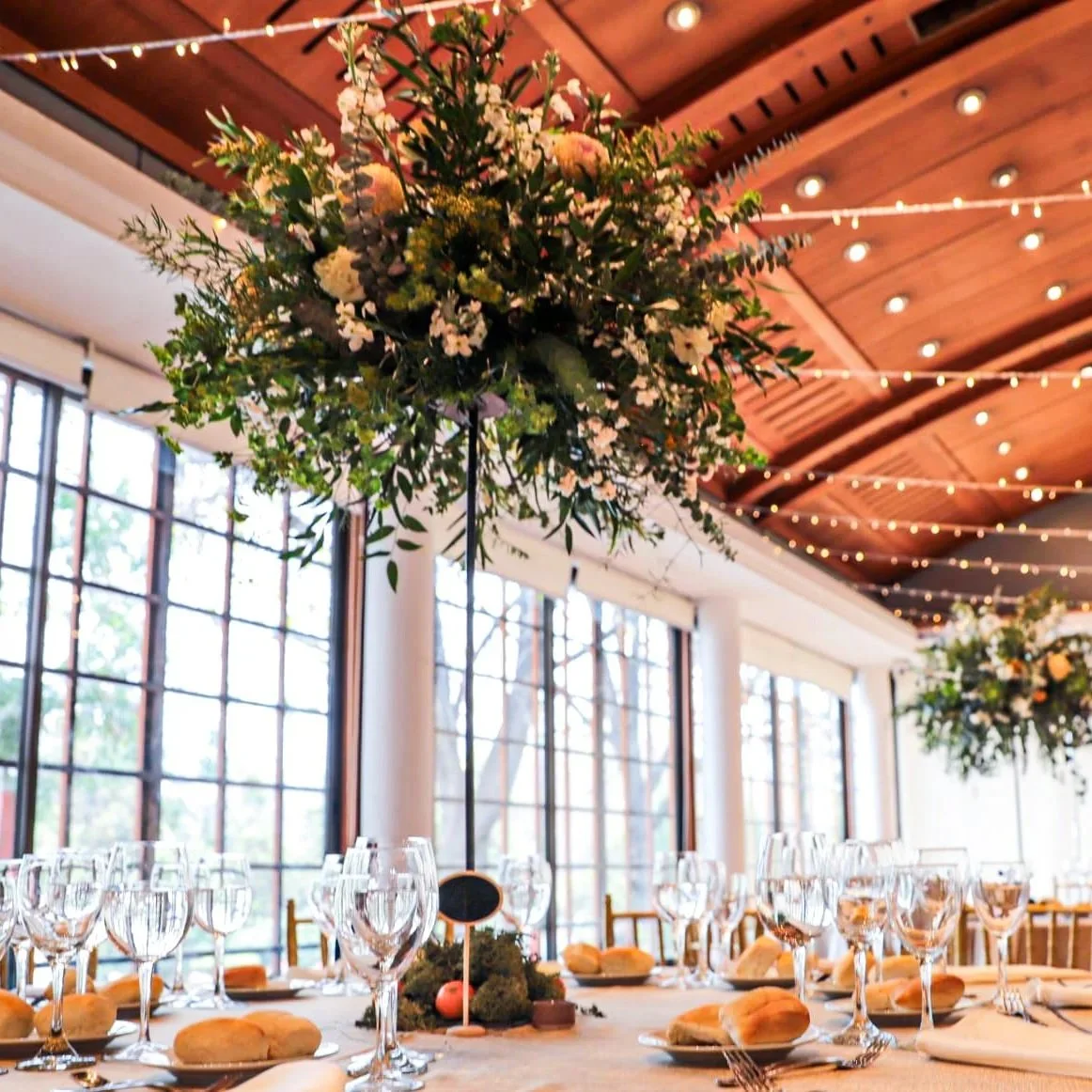 Mesas decoradas con copas de vino, panes y manteles blancos, en un espacio con paredes de ventanas grandes y un techo de madera, decorado con arreglos florales suspendidos y luces.
