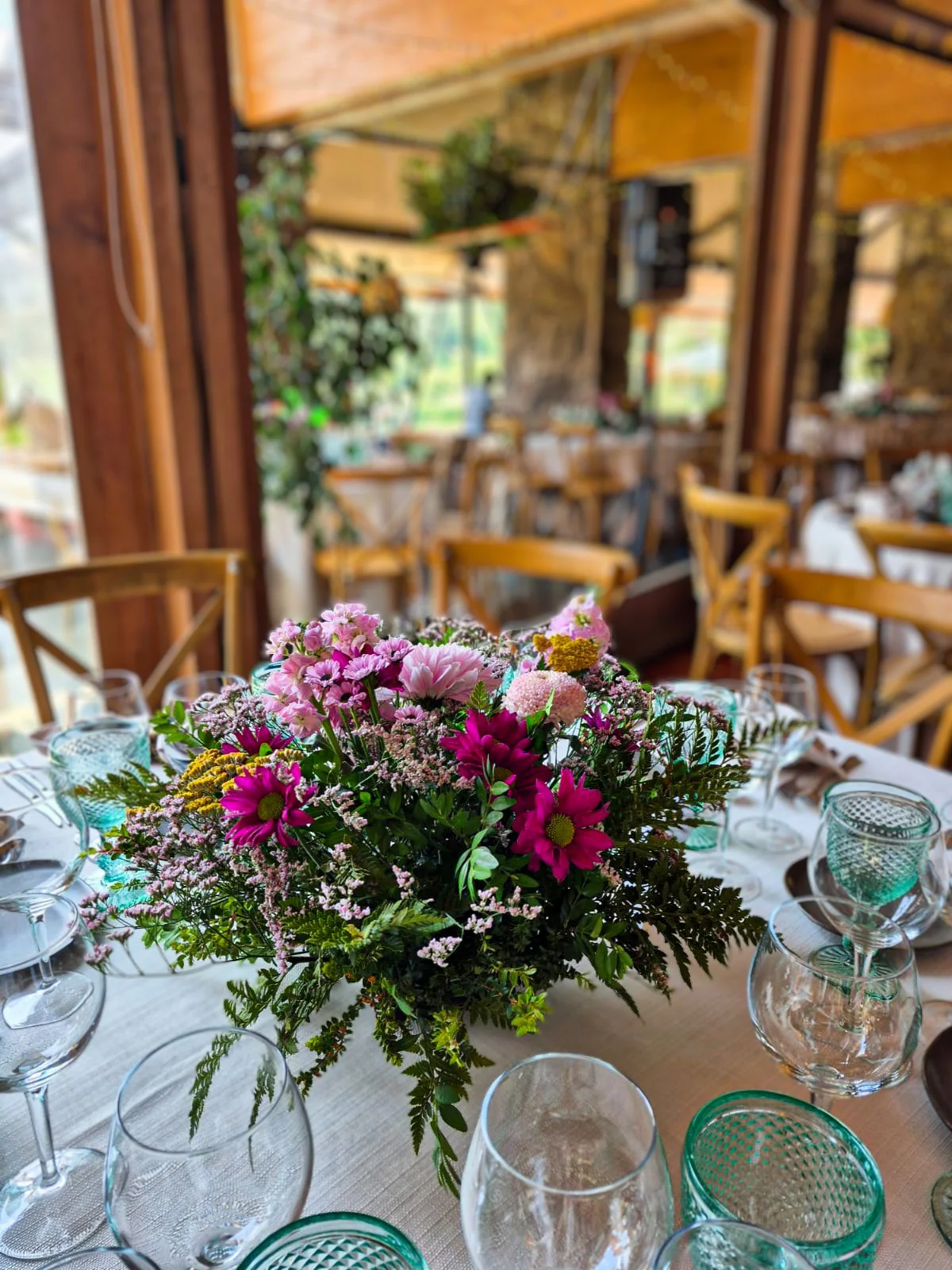 Centro de mesa con flores rosadas y moradas en una mesa decorada para un evento en un restaurante de madera y ventanas con vista al exterior.