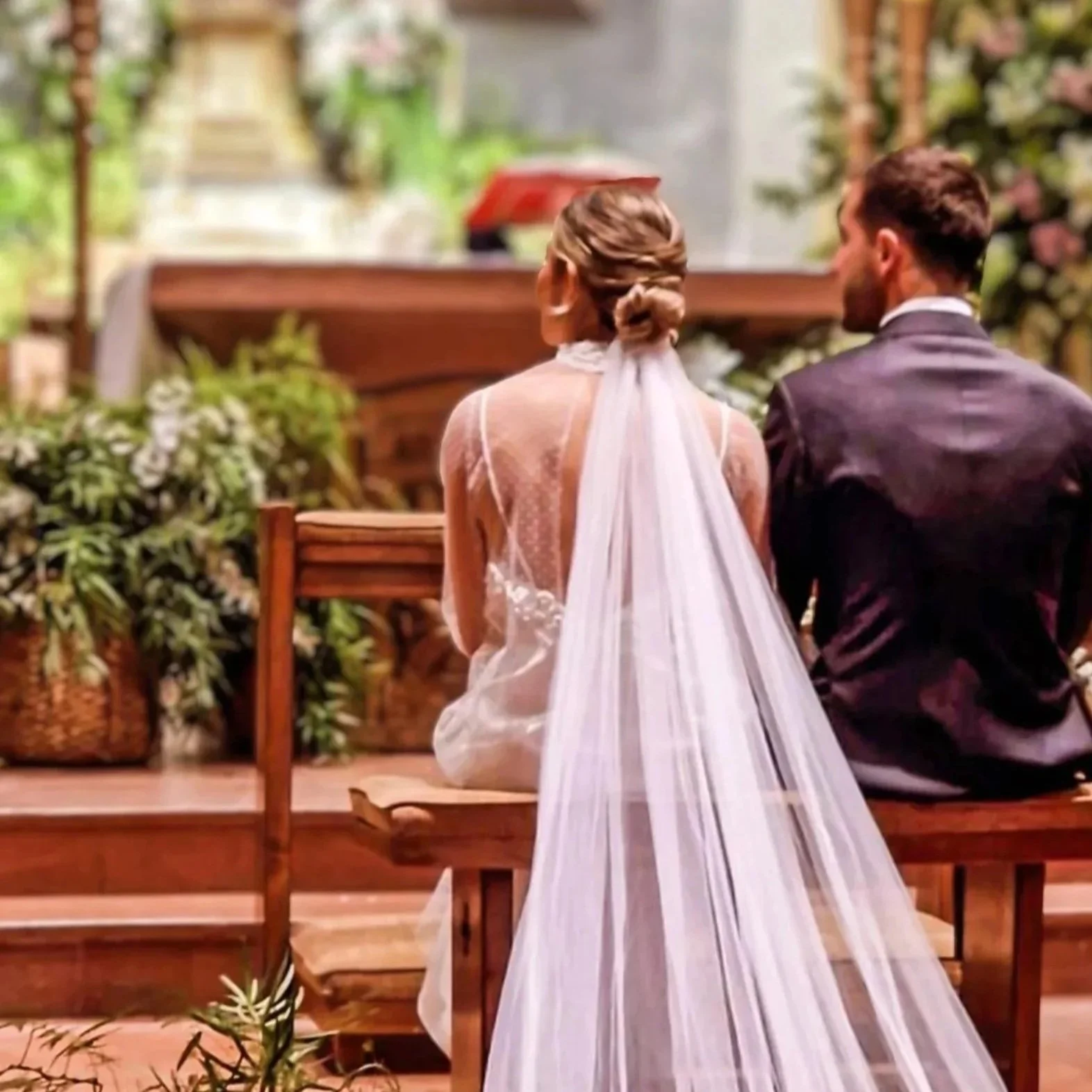 Una pareja en boda, sentados en un banco de madera en una ceremonia religiosa, ambos de Espaldas, ella con vestido de novia y él con traje oscuro, rodeados de plantas y flores, en un ambiente interior decorado.
