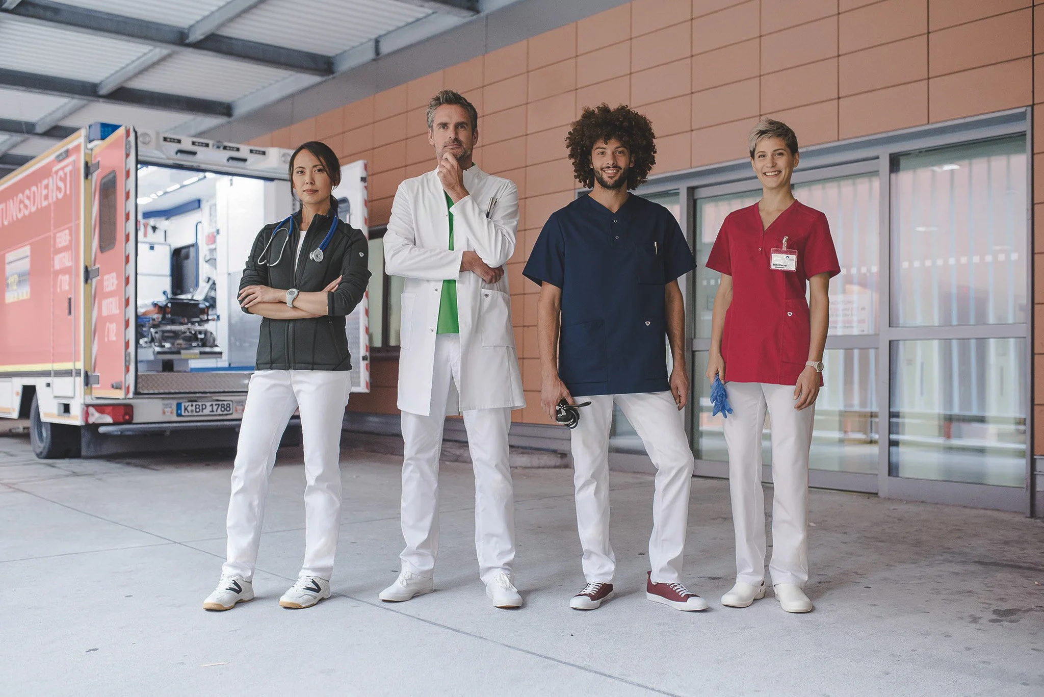 Quatre professionnels médicales debout devant un centre médical, avec une ambulance en arrière-plan, toutes souriantes et portant différentes uniformes.
