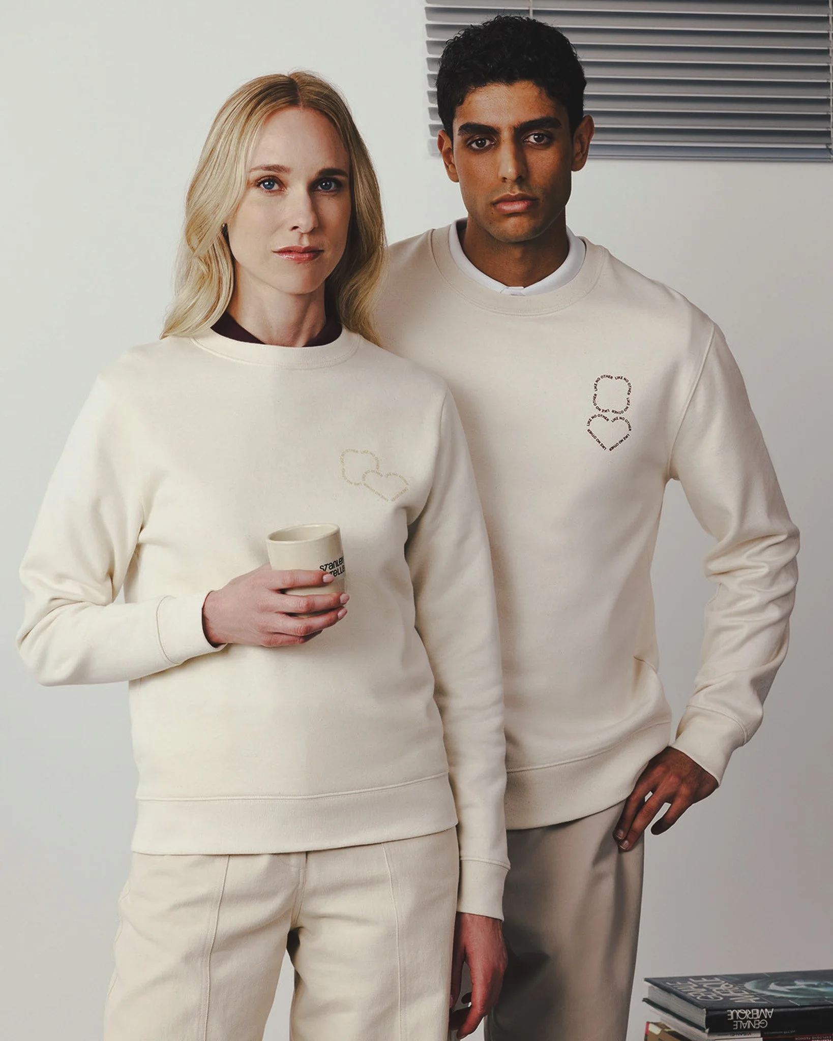 Deux personnes portent des sweats crème, l'une une tasse à café, dans un cadre intérieur minimaliste.