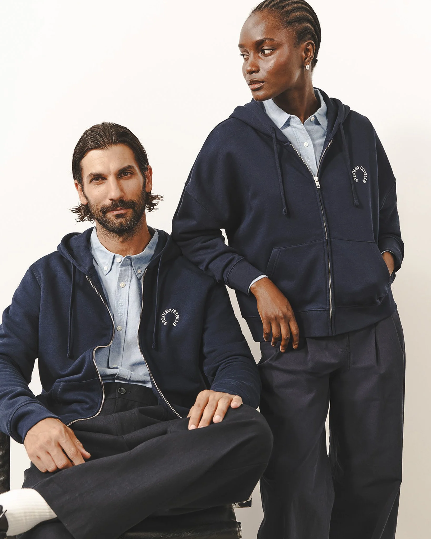 Deux personnes portent des sweats à capuche marines avec un logo blanc. L'une est assise, l'autre est debout à côté, dans un cadre neutre.
