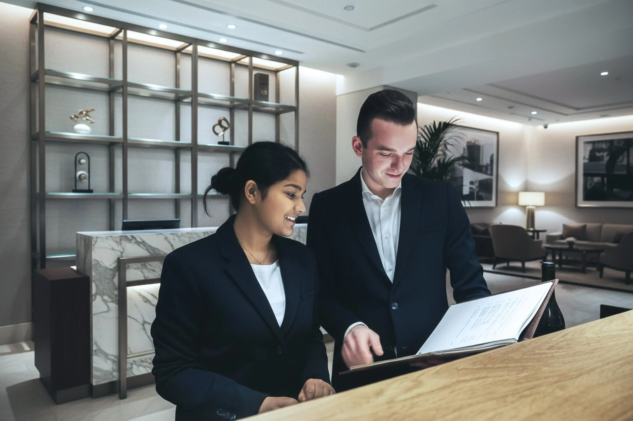 Deux personnes en tenue professionnelle, une femme et un homme, consultent un menu ou un document dans un lobby d'hôtel ou un espace d'accueil élégant.
