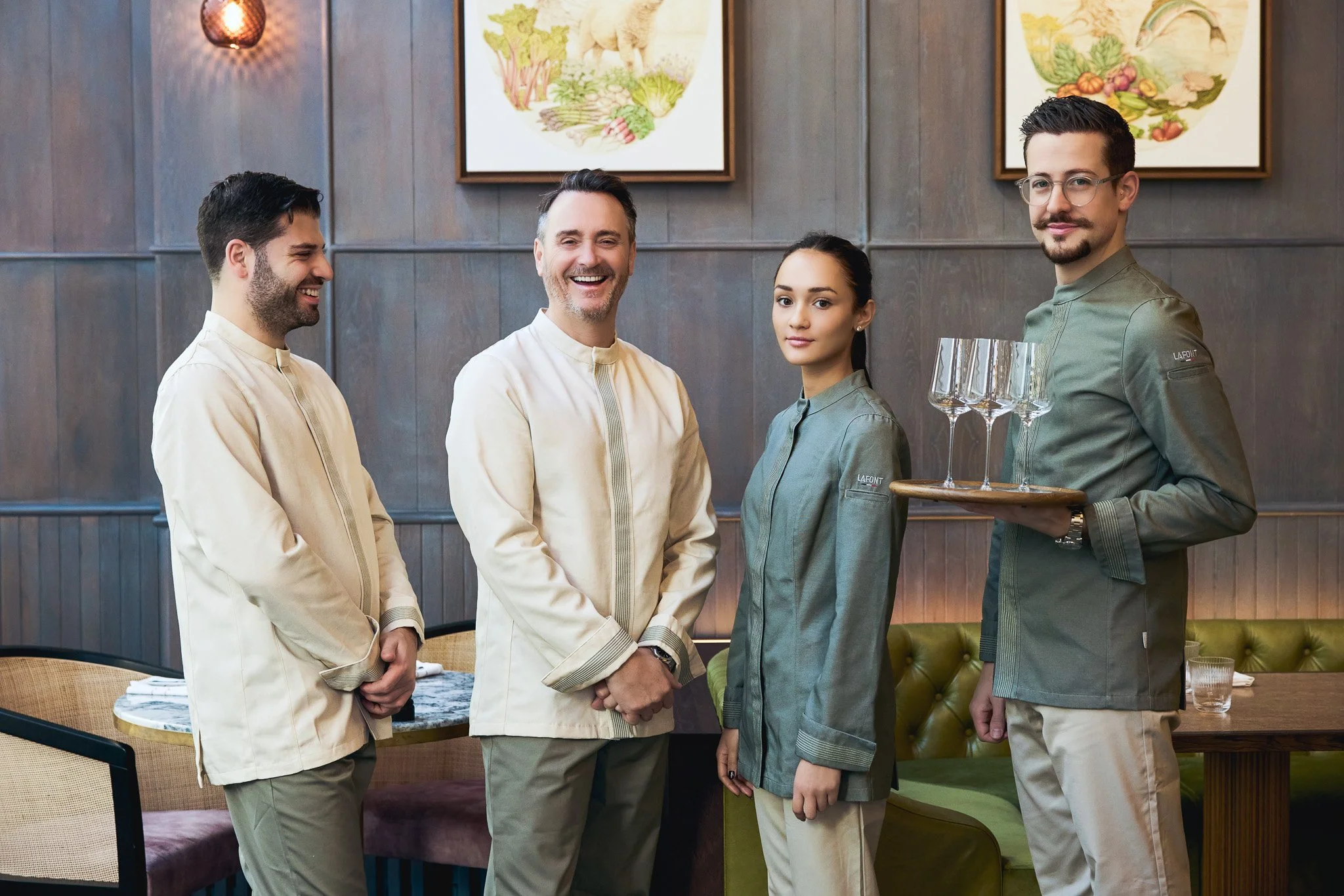 Quatre serveurs posant pour une photo dans un restaurant, deux hommes et une femme au centre, un autre homme à droite. L'homme de droite porte un plateau avec trois verres à vin. Le décor du restaurant comprend des tableaux avec des illustrations de légumes et d'animaux, et un mur en bois.