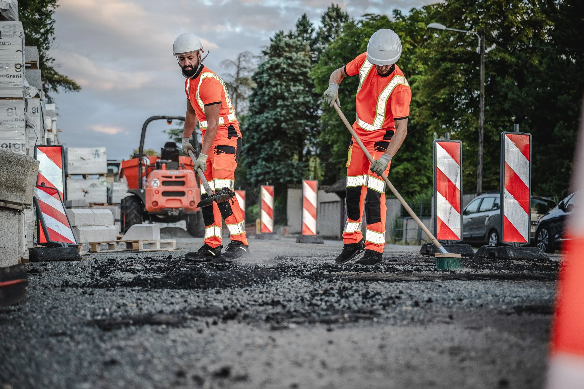 Deux ouvriers en tenue de sécurité orange travaillant sur une route en construction, utilisant des outils pour réparer la surface. En arrière-plan, des barrières de signalisation rouge et blanc et un arbre.