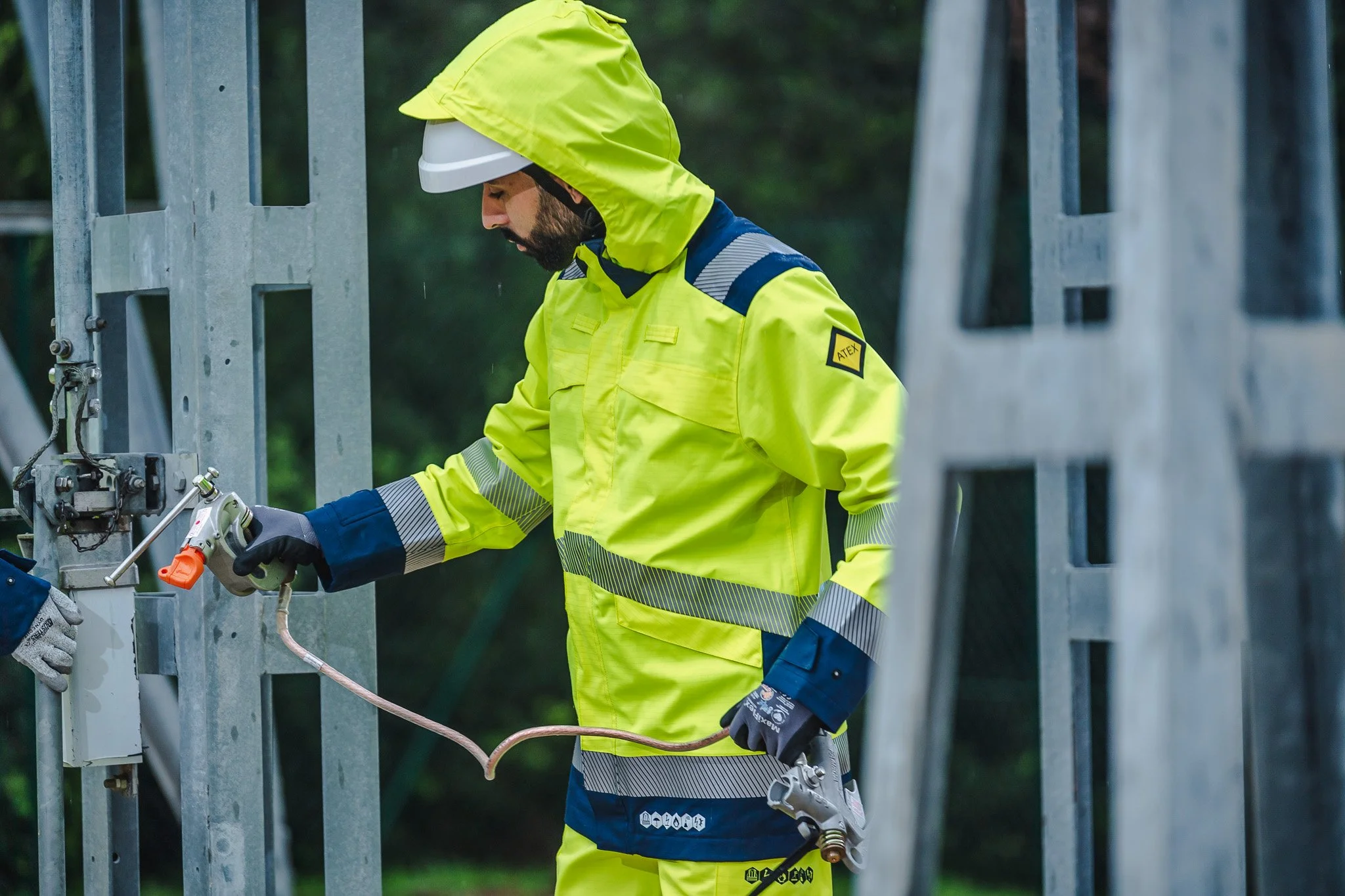 Un homme portant un équipement de sécurité jaune vif, un casque blanc, et des gants travaille sur une structure métallique lors d'une journée pluvieuse.