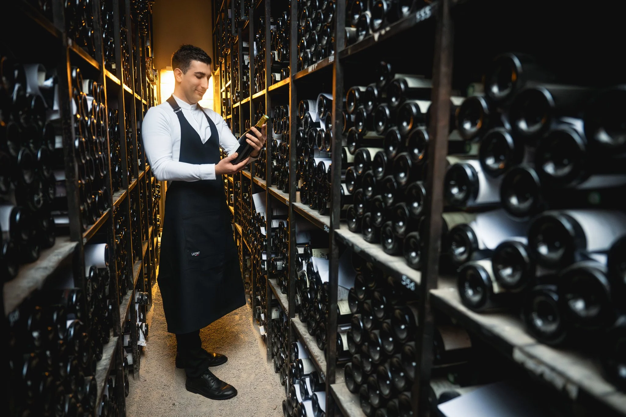 Un homme en uniforme noir et blanc, dans une cave à vin, regarde une bouteille de vin tout en tenant une autre bouteille dans sa main.