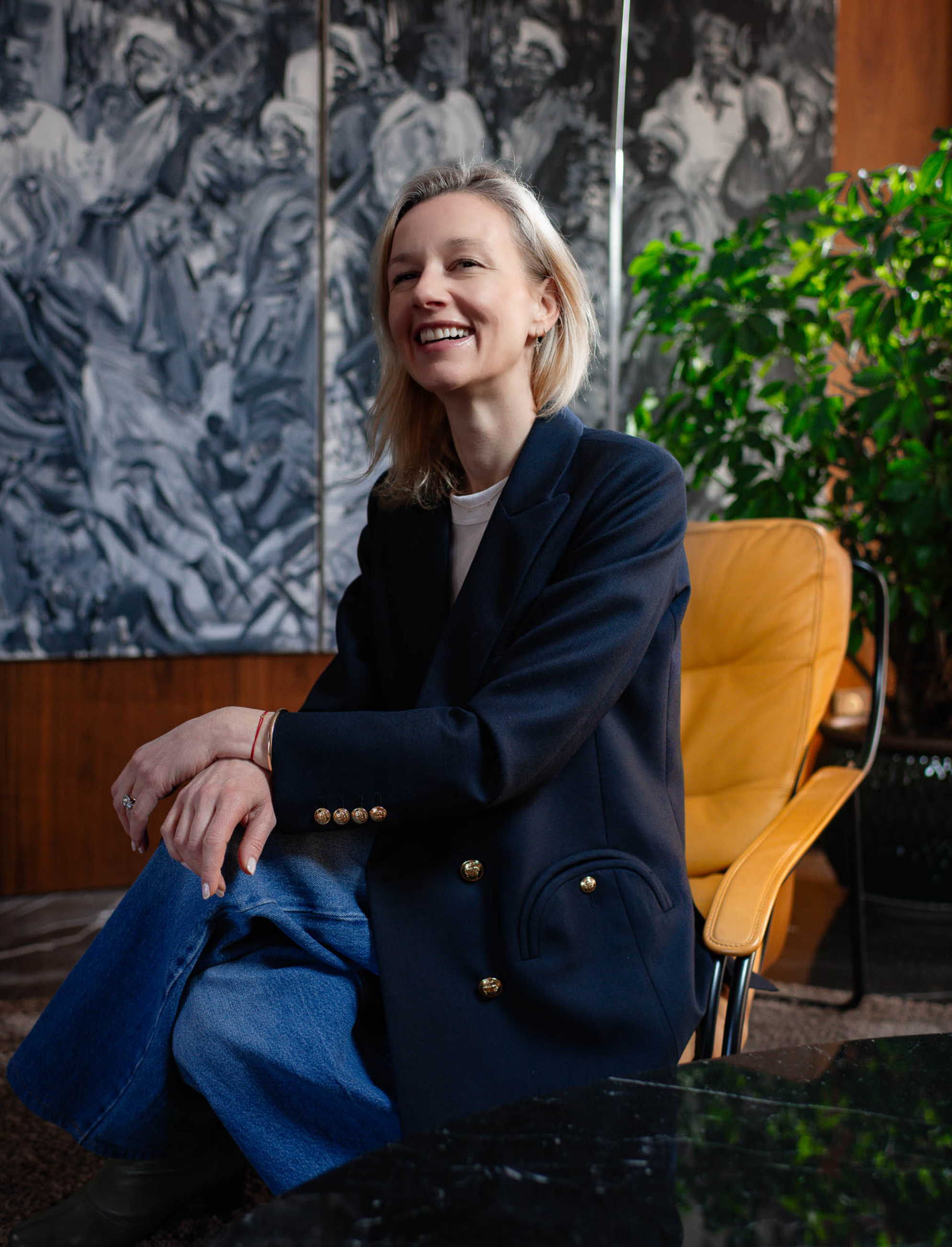 Une femme souriante assise sur une chaise en bois ou en cuir jaune, portant une veste noire à boutons dorés et un t-shirt blanc, dans un environnement intérieur avec une grande peinture murale en noir et blanc en arrière-plan et un poussin vert feuillage à sa droite.