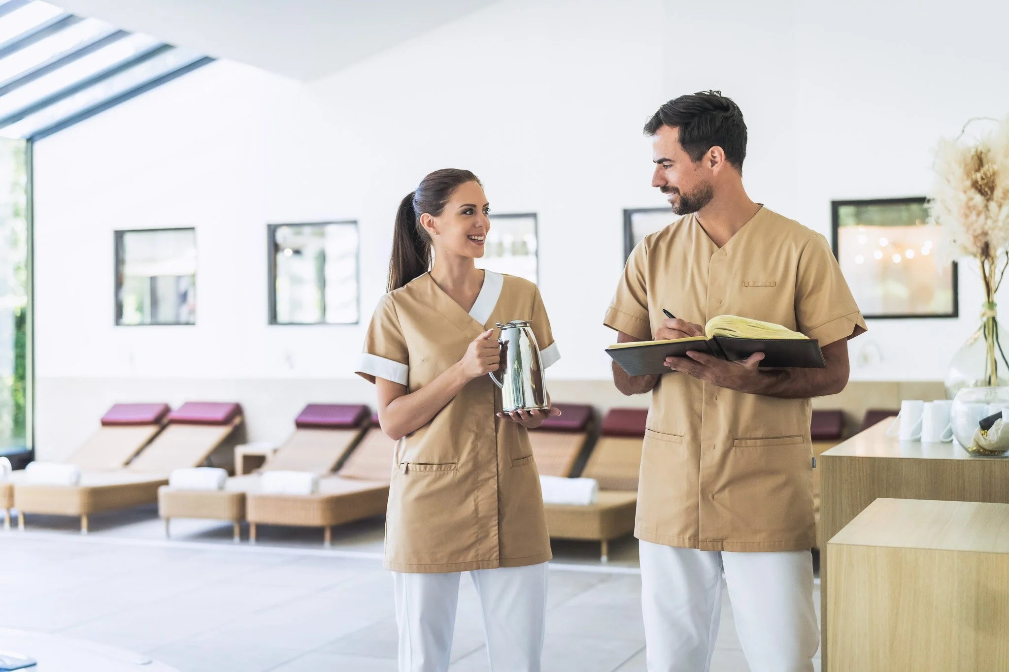 Deux employés en uniformes de spa ou centre de bien-être discutent dans une salle lumineuse et moderne, avec des chaises longues en arrière-plan.