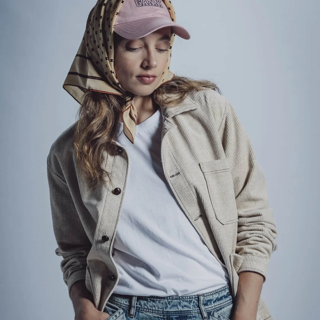 Une jeune femme porte un bonnet rose, une écharpe beige à pois, une veste beige en velours côtelé, un t-shirt blanc et un jean. Elle a les yeux fermés et un petit sourire.