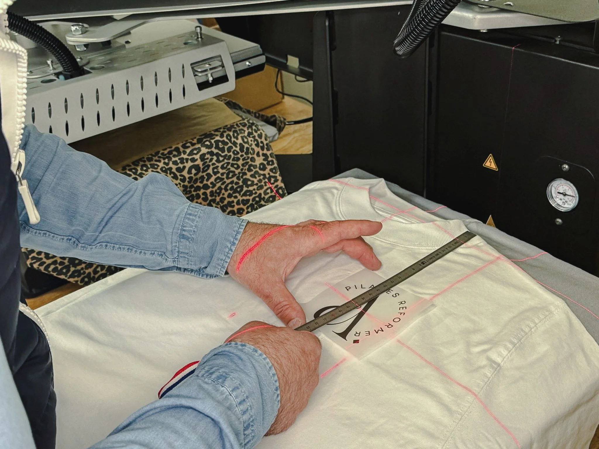 Une main tient une règle à seuil sur un tissu blanc avec des lignes rouges, probablement pour une couture, dans un espace de travail lié à la mode ou à la couture.