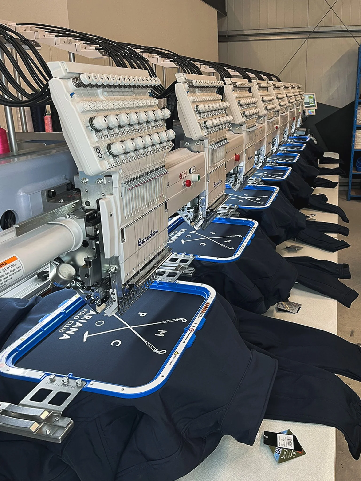 Une rangée de machines à coudre industrielles avec des fils de différentes couleurs et des tissus noirs, apparemment utilisées pour la fabrication de vêtements ou d'articles textiles.