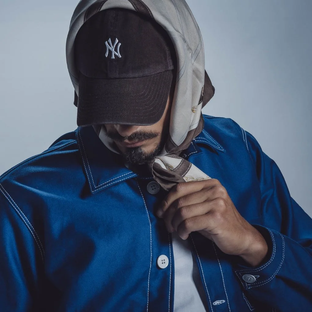 Un homme portant une casquette noire avec un logo de Yankees de New York, un foulard beige, une veste en denim bleu avec des points de couture contrastants, et une chemise blanche, regardant vers le bas