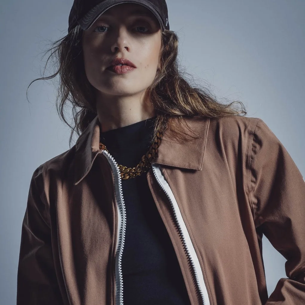 Une femme porte une casquette noire, un manteau marron et un collier en or, avec un fond gris.