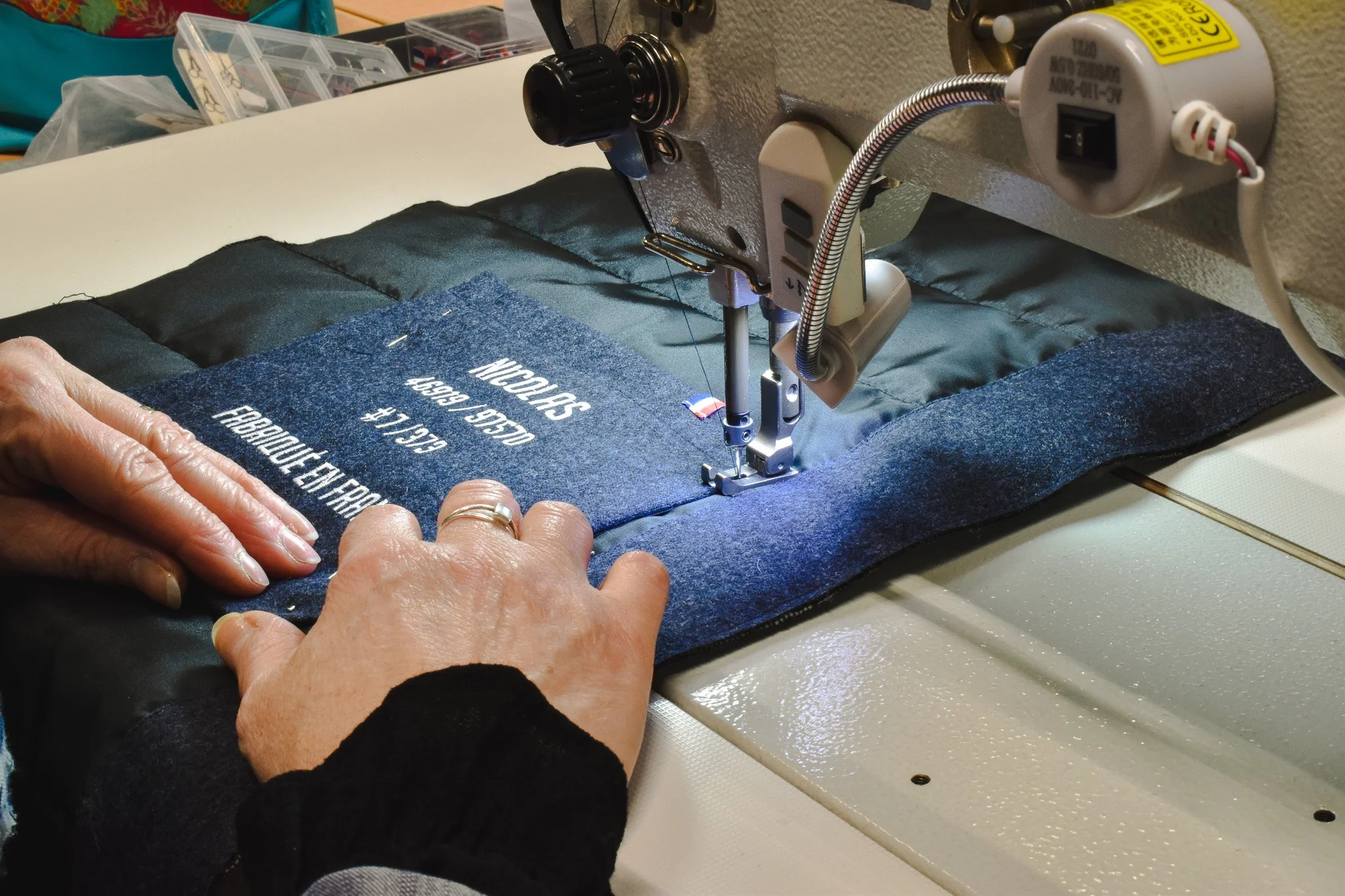 Une personne utilise une machine à coudre pour assembler une pièce de tissu bleu foncé avec des textes en blanc. La machine est fixée sur une table, avec des mains guidant le tissu lors de la couture.