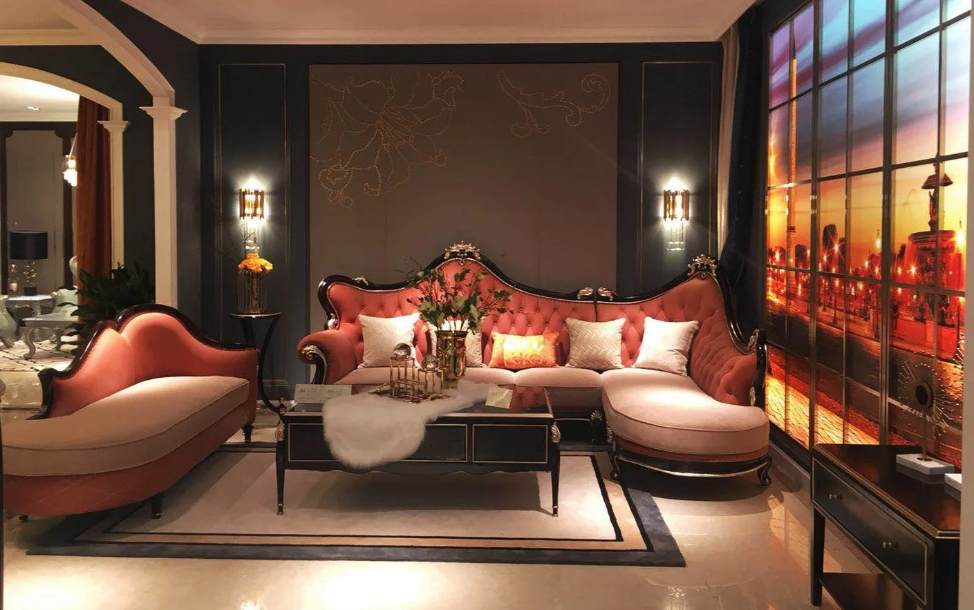 Elegant living room with a vintage pink velvet sofa, matching chaise lounge, and decorative pillows. A central black wooden coffee table with a white fur runner, floral arrangement, and small decorative items. The room features dark walls with wall sconces, a large window with a cityscape sunset view, and elegant curtains on the side. Additional seating and decor are visible in the background.