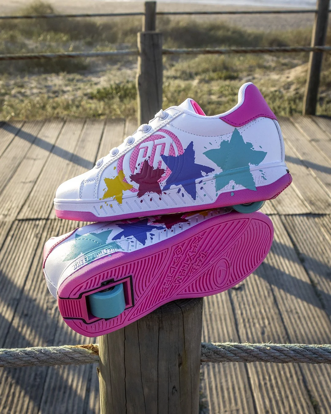 A colorful pair of roller skates with pink, blue, yellow, and teal star designs, placed on a wooden post outdoors with a beach scenery in the background.