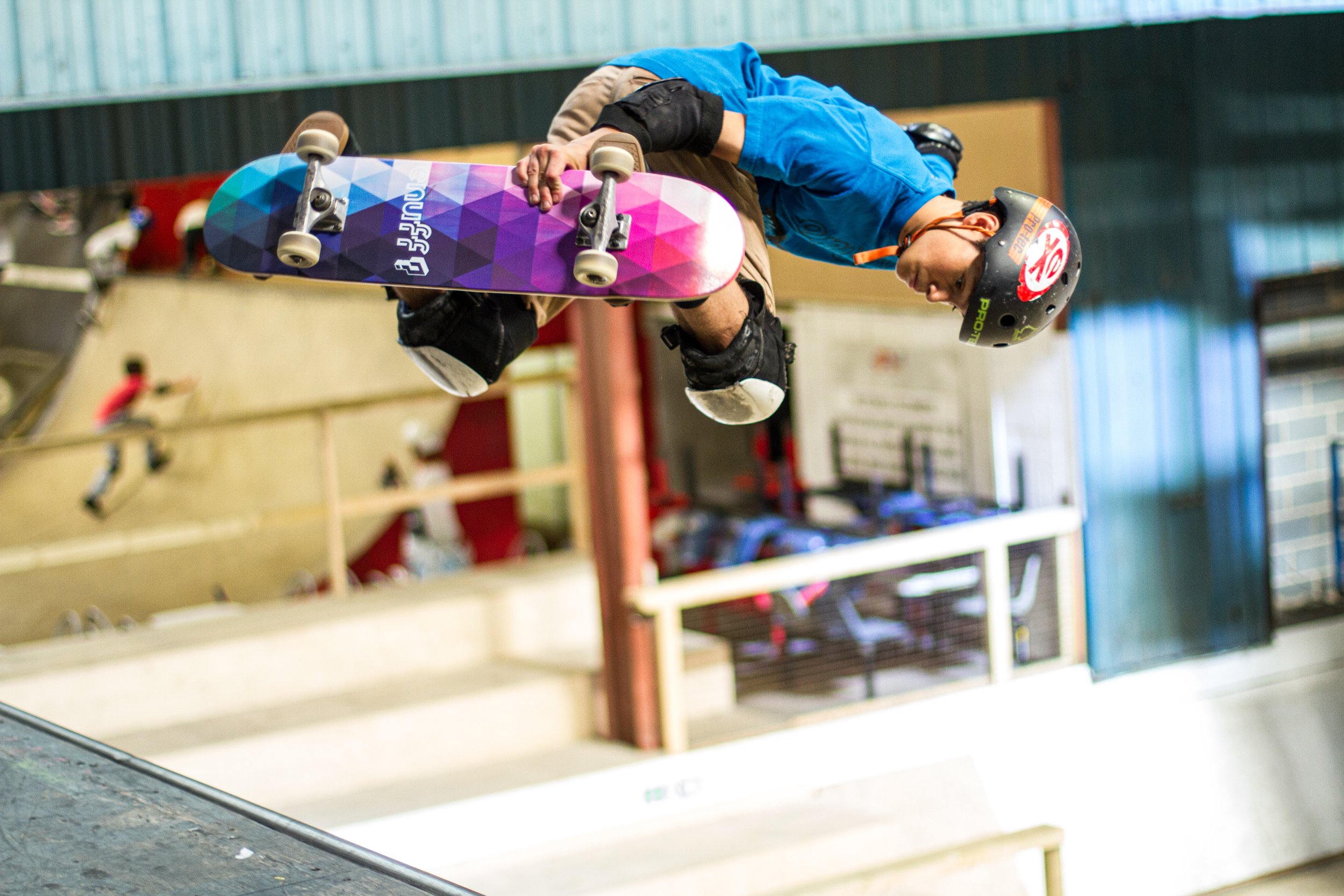 A person wearing a black helmet and protective gear is skateboarding at an indoor skatepark with a colorful skateboard and a blue shirt.