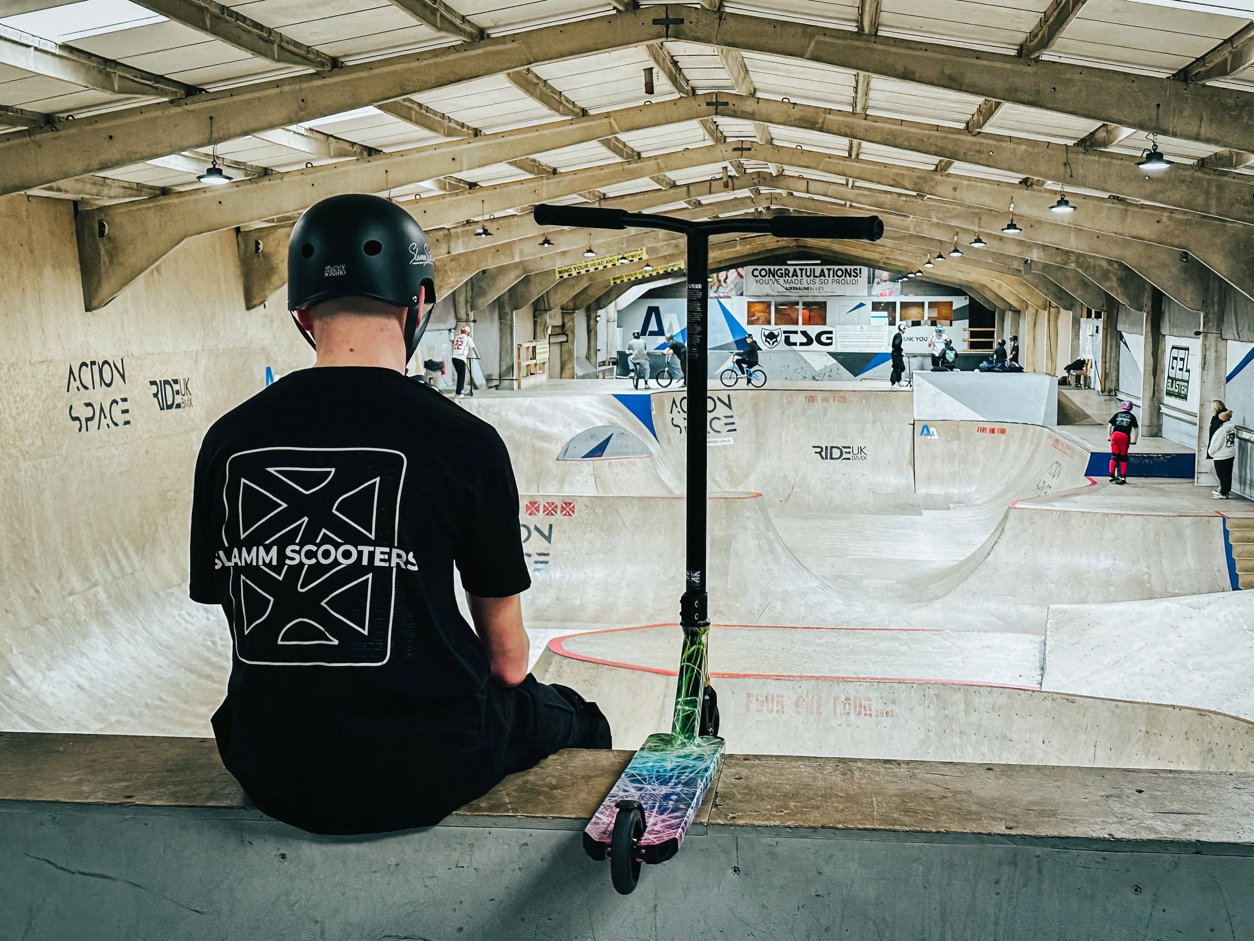 Interior of a skatepark with a young person wearing a helmet and sitting on a bench, holding a skateboard. In the background, there are multiple people riding bikes and skating on ramps.