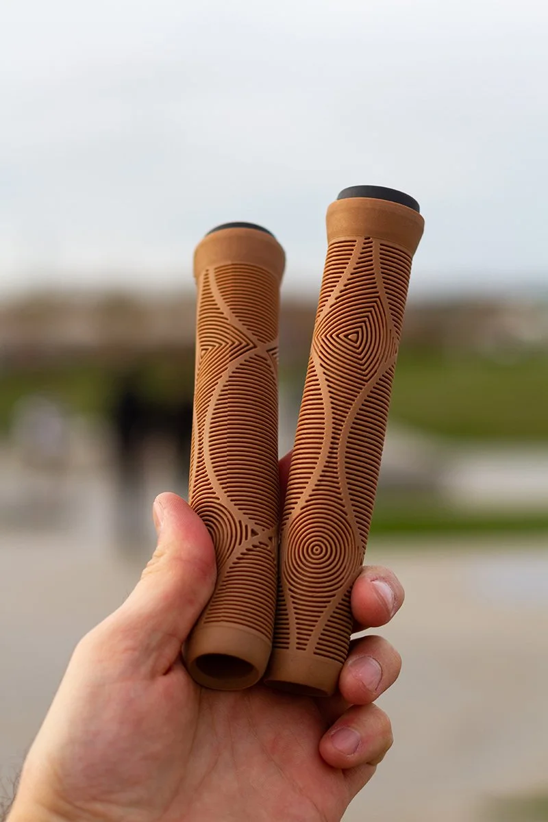 Close-up of two brown rubber bicycle handlebar grips with textured geometric patterns, held in a person's hand outdoors.