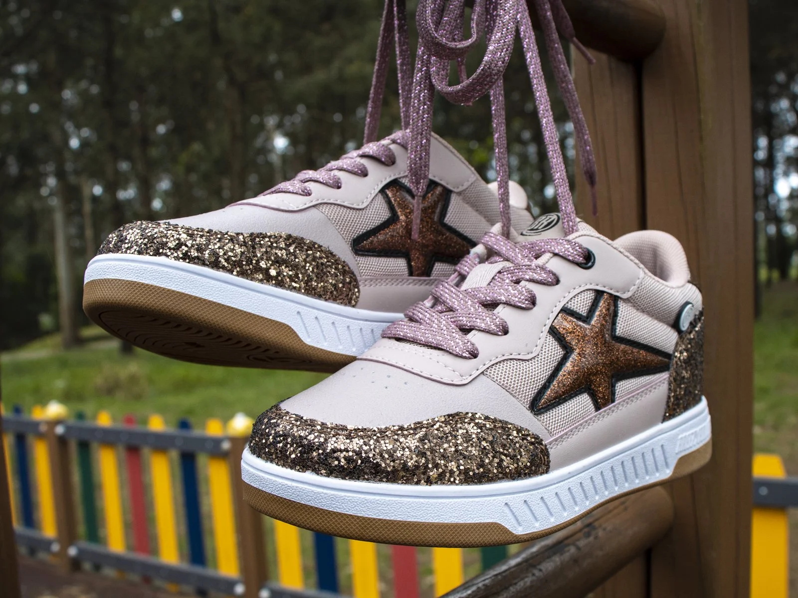 A pair of pink sneakers with glitter accents, star designs, pink shoelaces, and a white sole, hanging outdoors on a wooden structure.