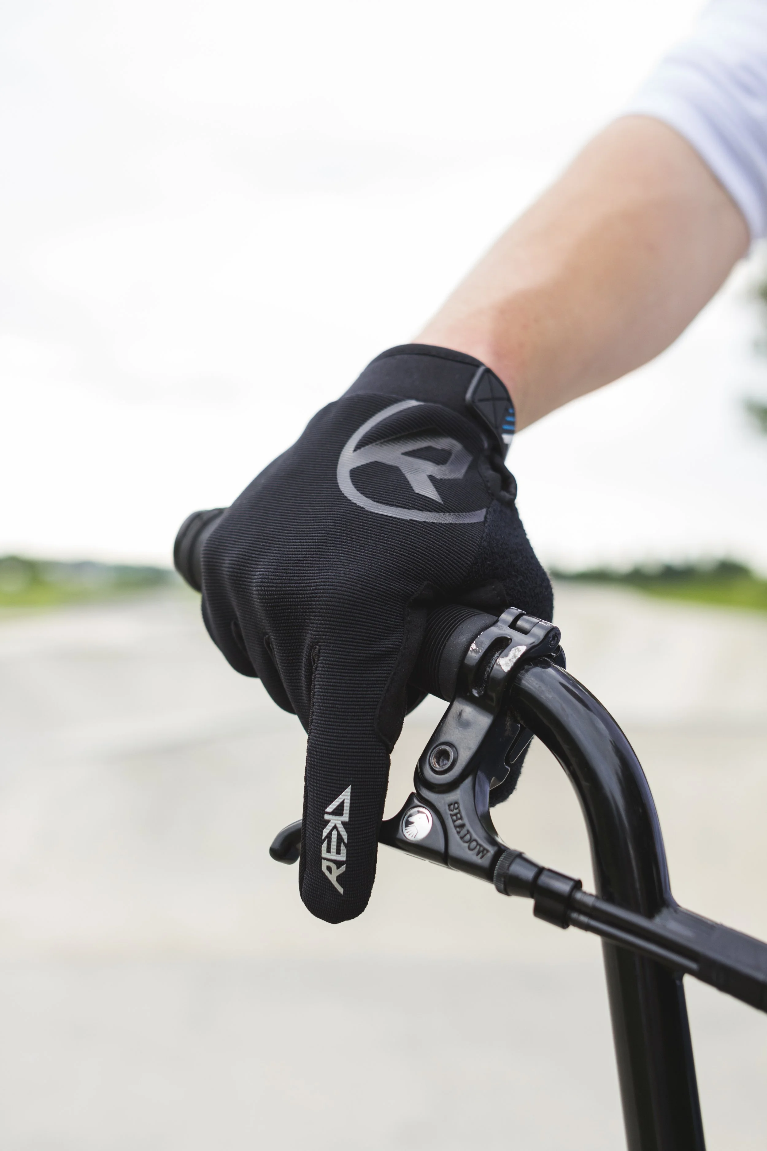 Close-up of a rider's hand gripping a mountain bike handlebar, wearing a black glove with the logo REI, with a blurred outdoor background.