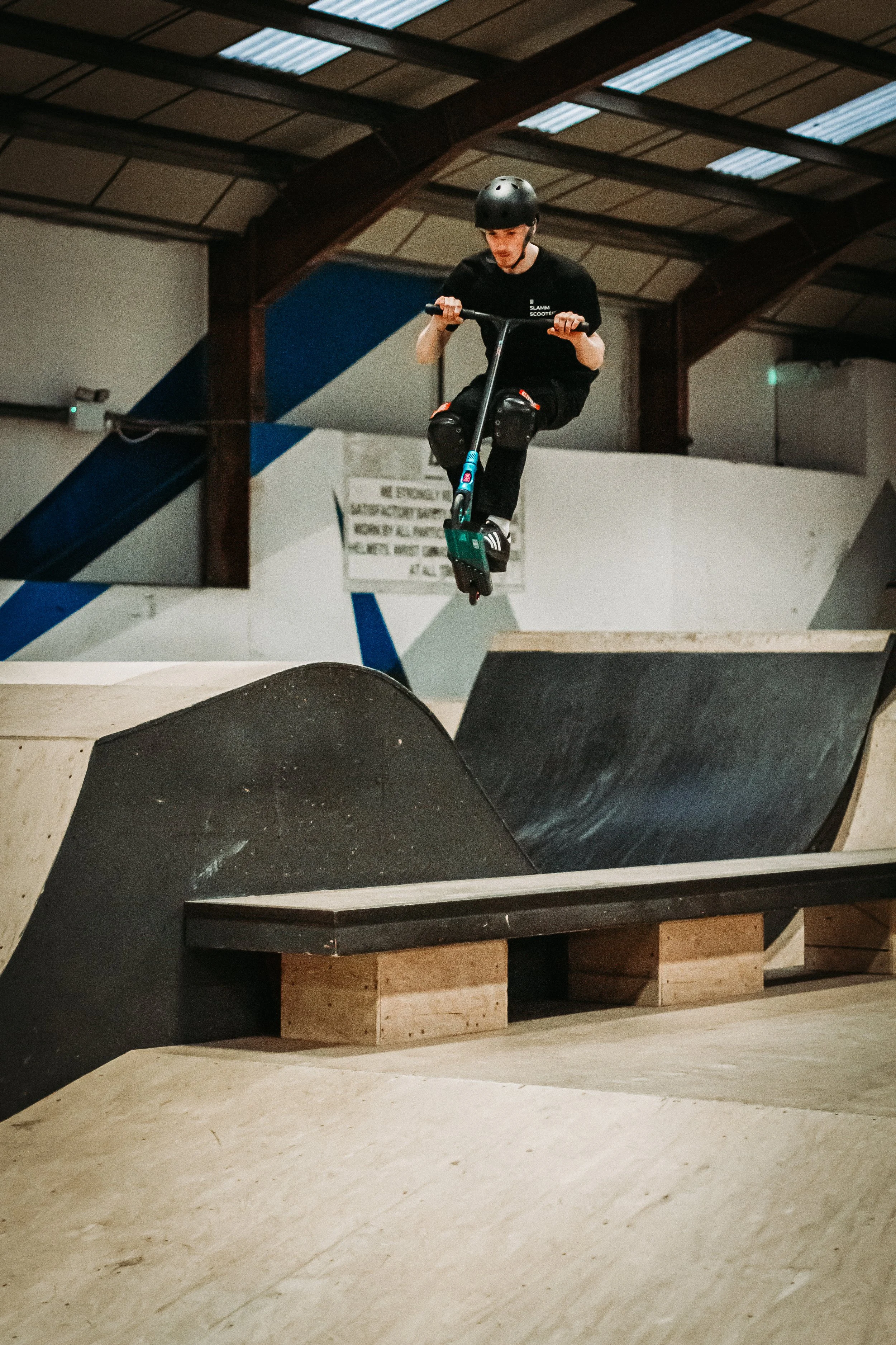 A person wearing a black helmet and black T-shirt is riding a scooter at an indoor skate park. The skate park has ramps and a wooden floor.