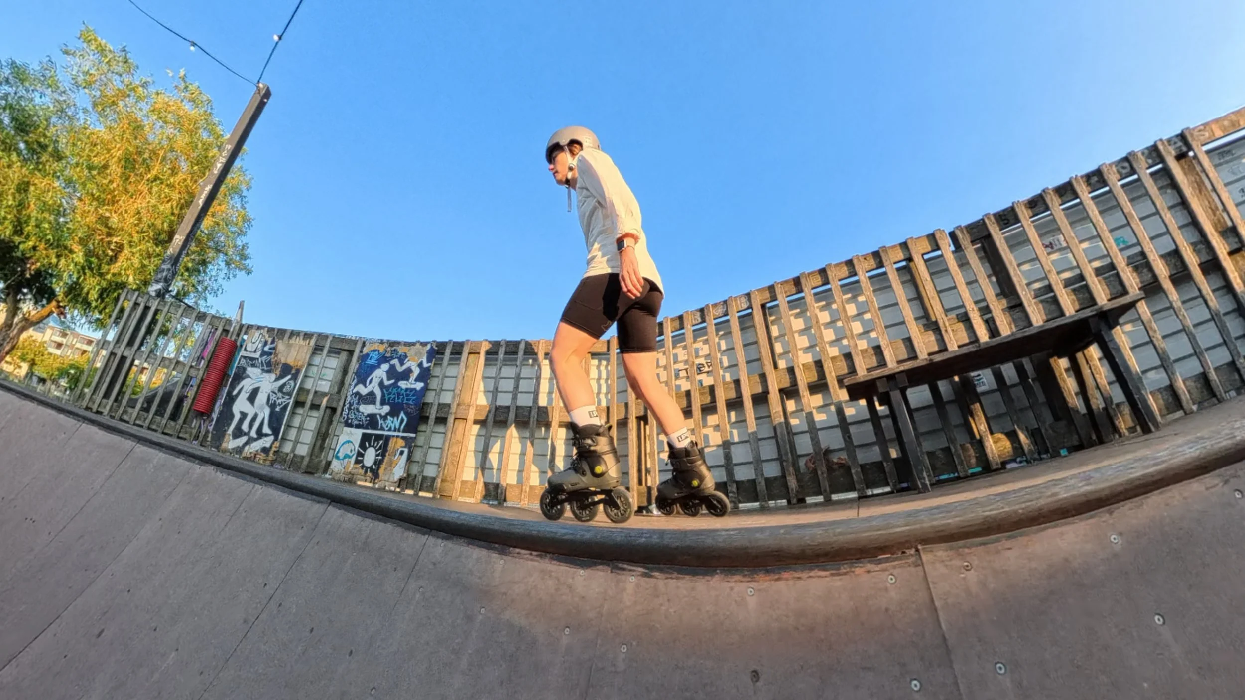 A person roller skating on a wooden skatepark ramp, wearing a helmet, sunglasses, shorts, a long-sleeve shirt, and protective gear. The background features a graffiti-covered fence, a tree with green and yellow leaves, and a clear blue sky.