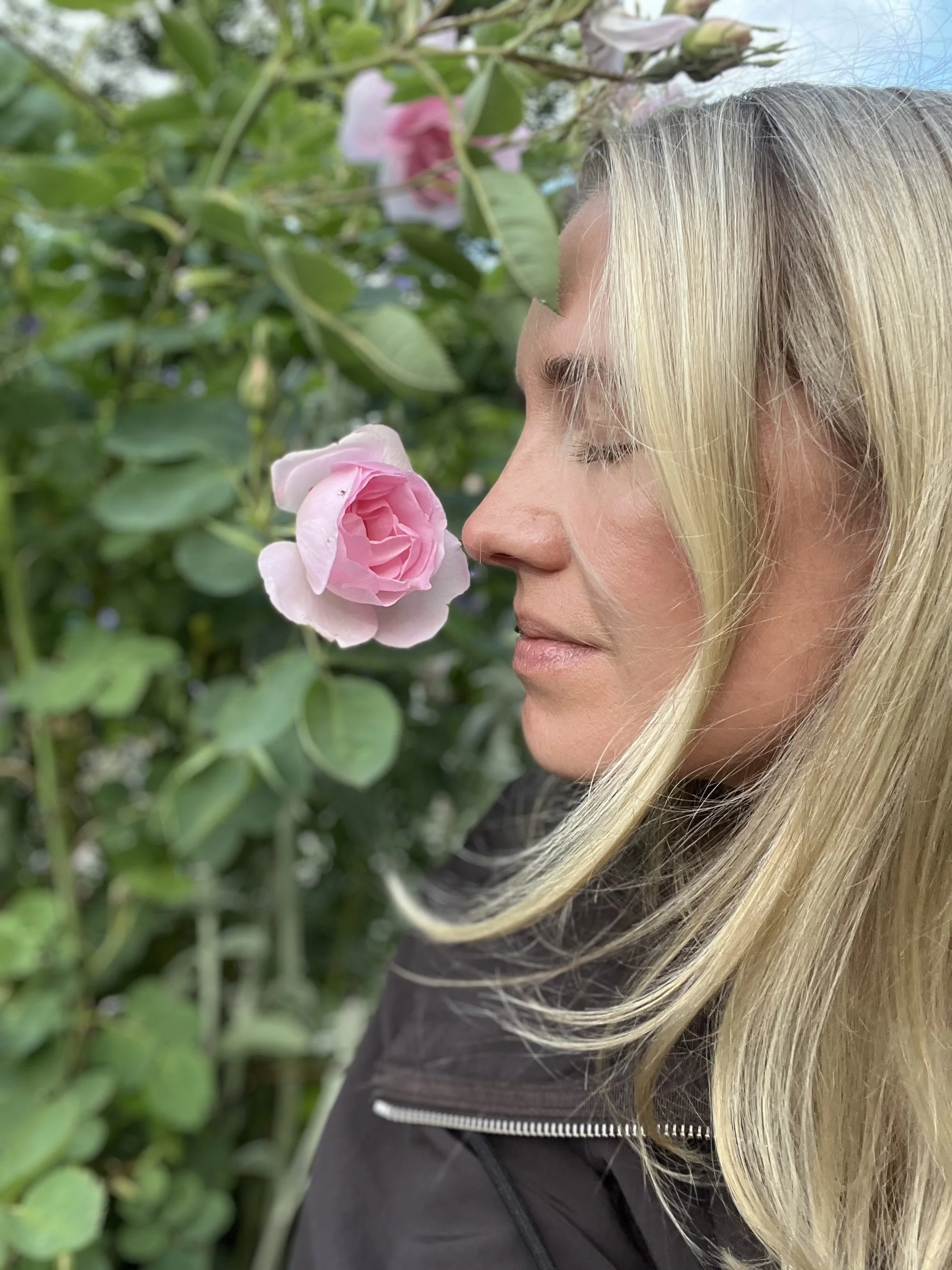 A woman with platinum blonde hair and light skin is depicted in profile with her eyes closed, facing a pink rose bush. The background is filled with green leaves and pink roses, and she is wearing a black jacket with an exposed zipper.