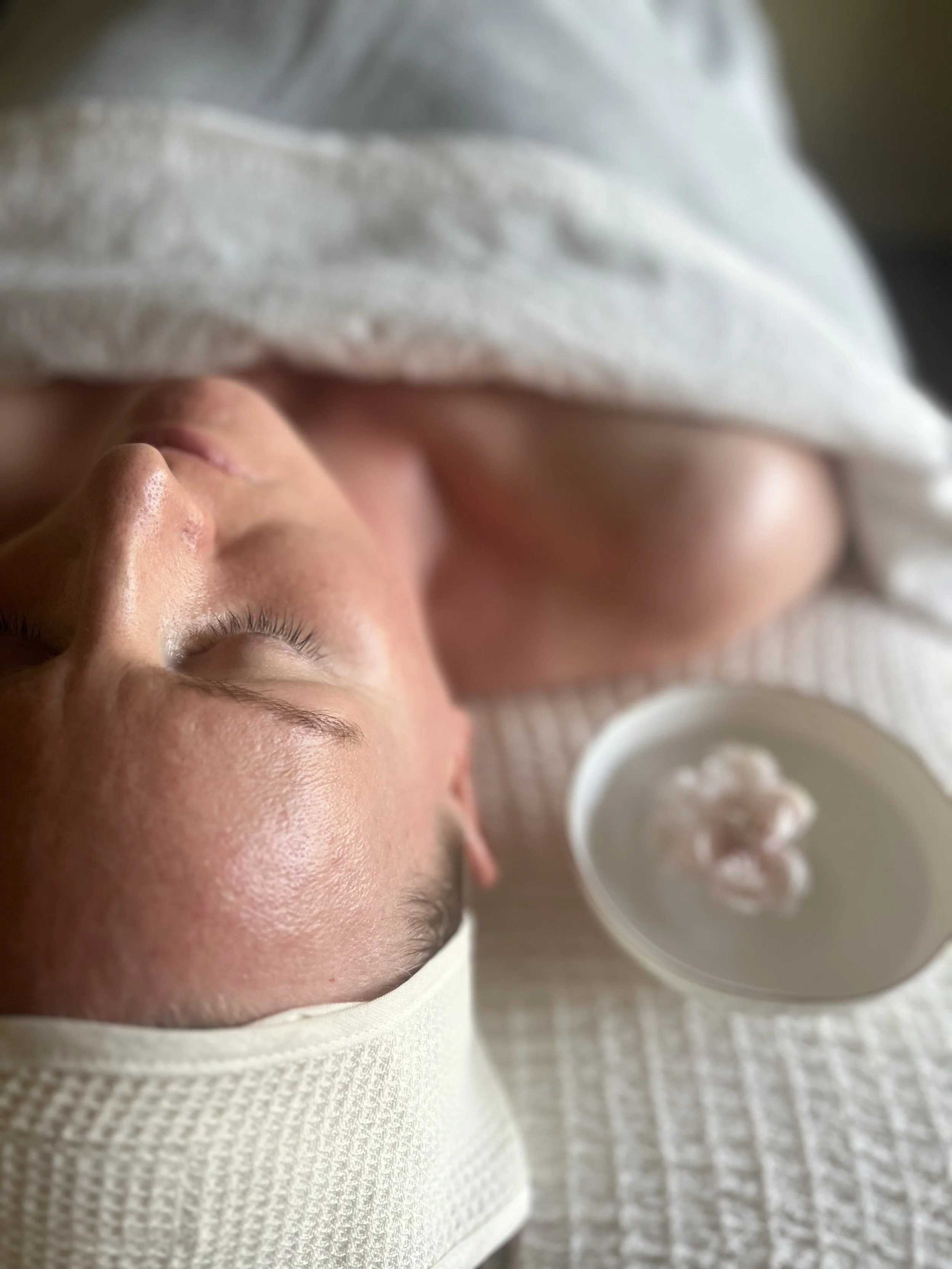 Close-up of a person's face with closed eyes, lying on a textured white surface, with a bowl of cream nearby.