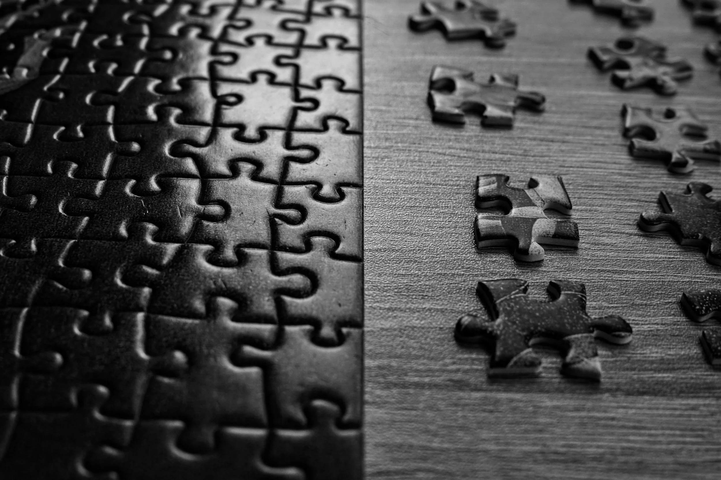 Black and white photo of jigsaw puzzle pieces, some assembled and some scattered on a wooden surface.