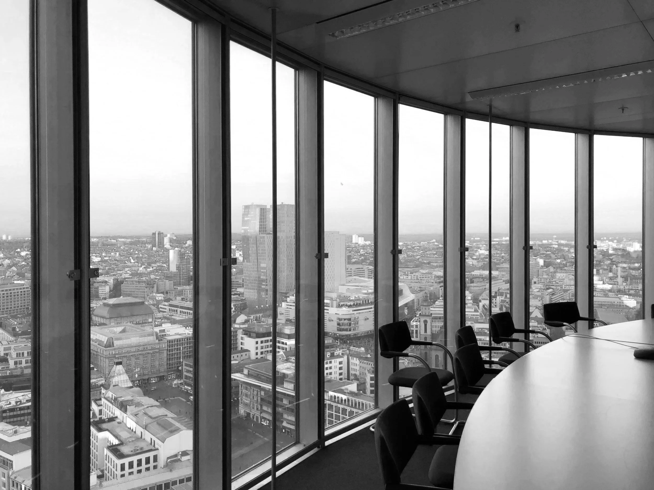 Black and white photo of a conference room with large floor-to-ceiling windows showing a city skyline with tall buildings.