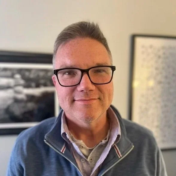 A man with glasses and short hair smiling indoors with blurred framed pictures in the background.