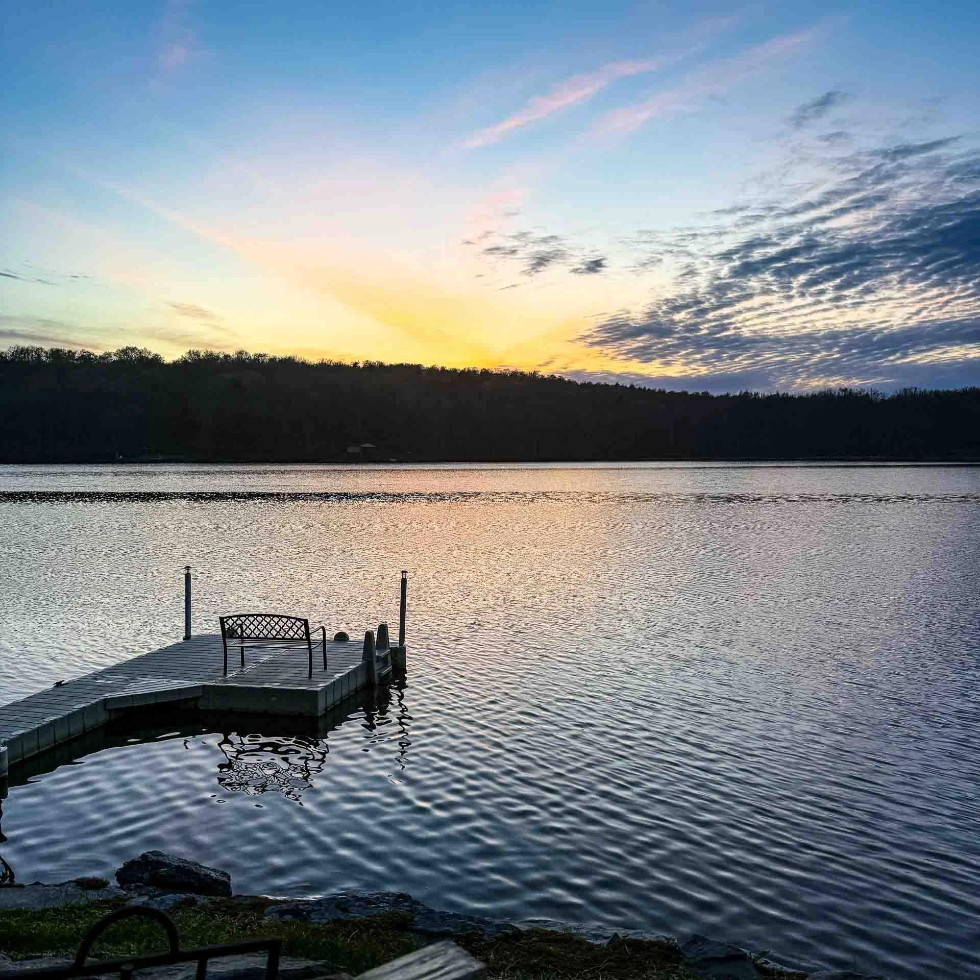 Sunset over a calm lake with a wooden dock and empty chairs, surrounded by trees.